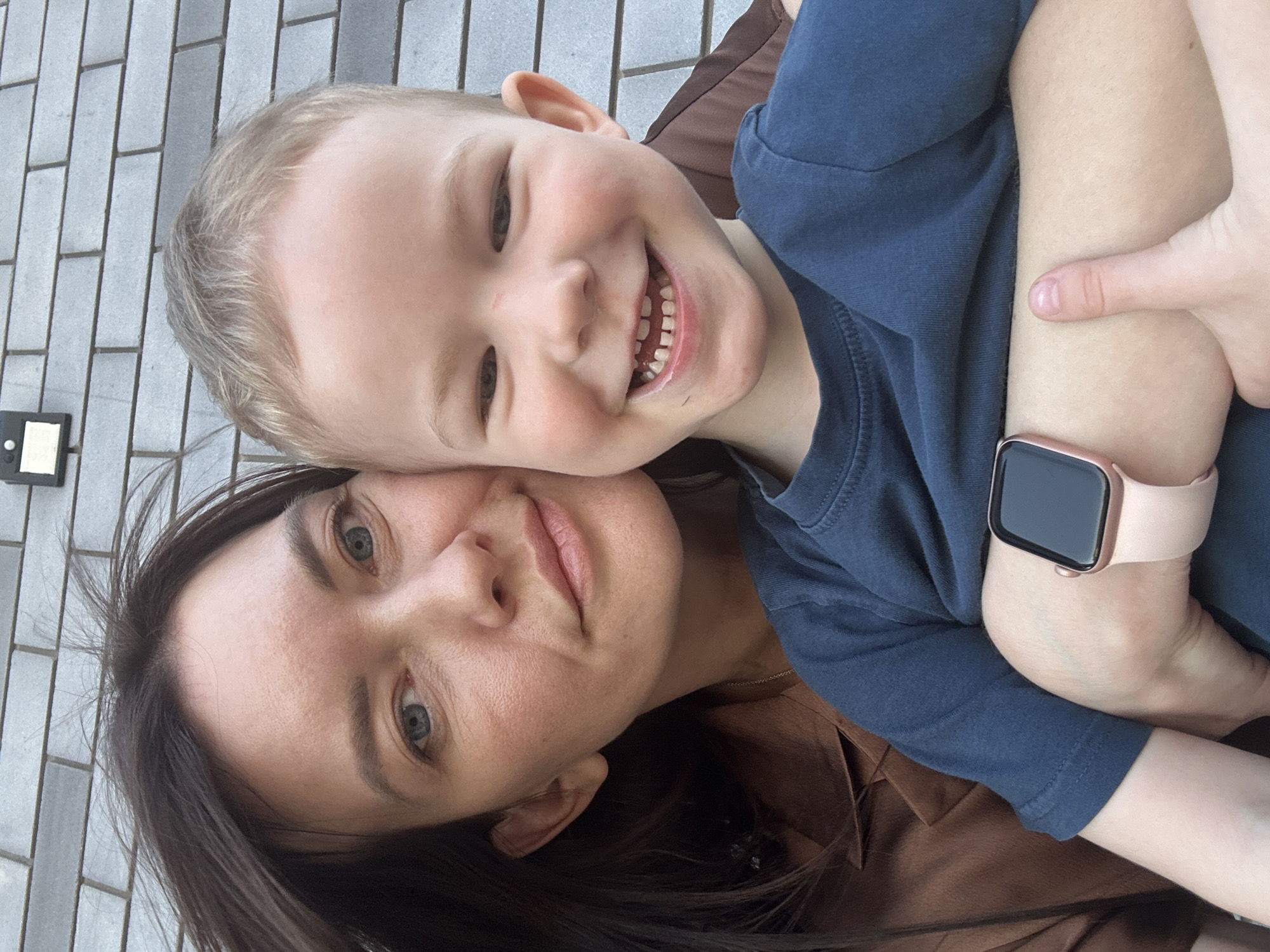 A young child and adult are posing closely for a photograph. The adult, a woman, has dark hair and is wearing a brown shirt. The child, presumably male, is wearing a dark blue shirt. The woman is holding the child, his arms around her, as they face the camera. The woman is wearing a smart watch on her wrist. The location is identified as Прикамский, Russia, though no specific geographical details are readily apparent in the image. The background includes a gray brick surface.