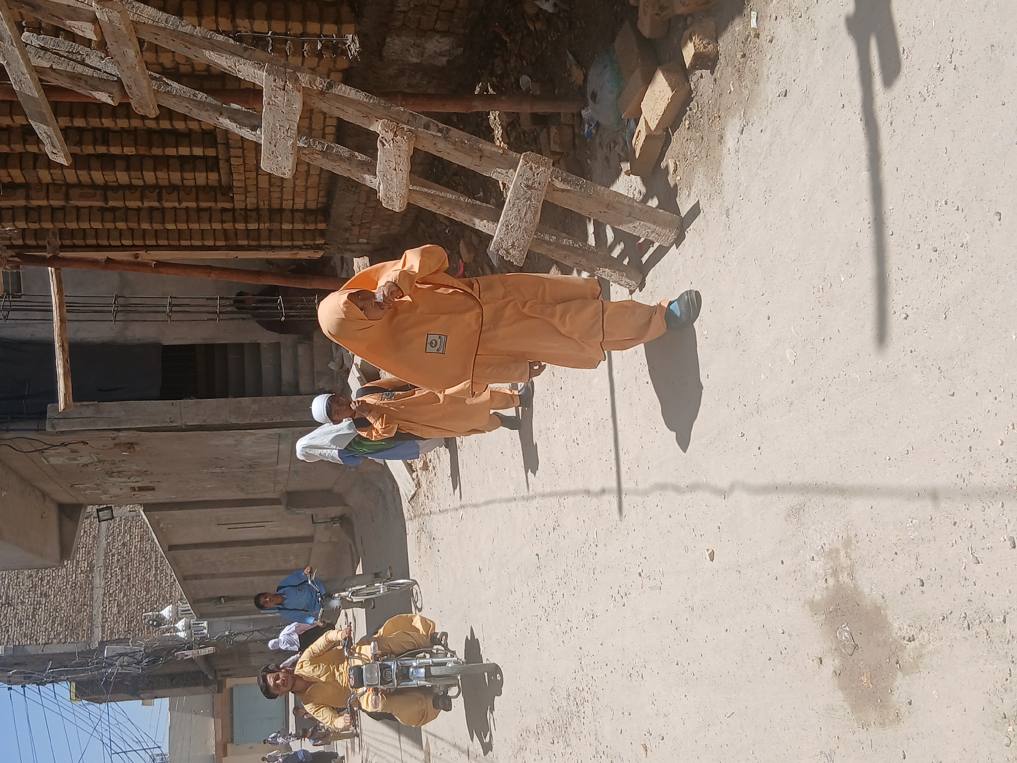 The image depicts a street scene in Quetta City Tehsil, Pakistan, under bright sunlight. The street appears to be dusty or unpaved. In the foreground, a person in an orange shalwar kameez is walking. Nearby, a boy wearing a white skullcap and light blue shalwar kameez stands facing the same direction. Further down the street, a person wearing a yellow shalwar kameez rides a motorbike and another person in a blue shirt and pants rides a bicycle. Overhead, there is a building under construction. Wooden beams and what appear to be unfinished brickwork indicate the ongoing construction. Discarded bricks and debris are scattered.