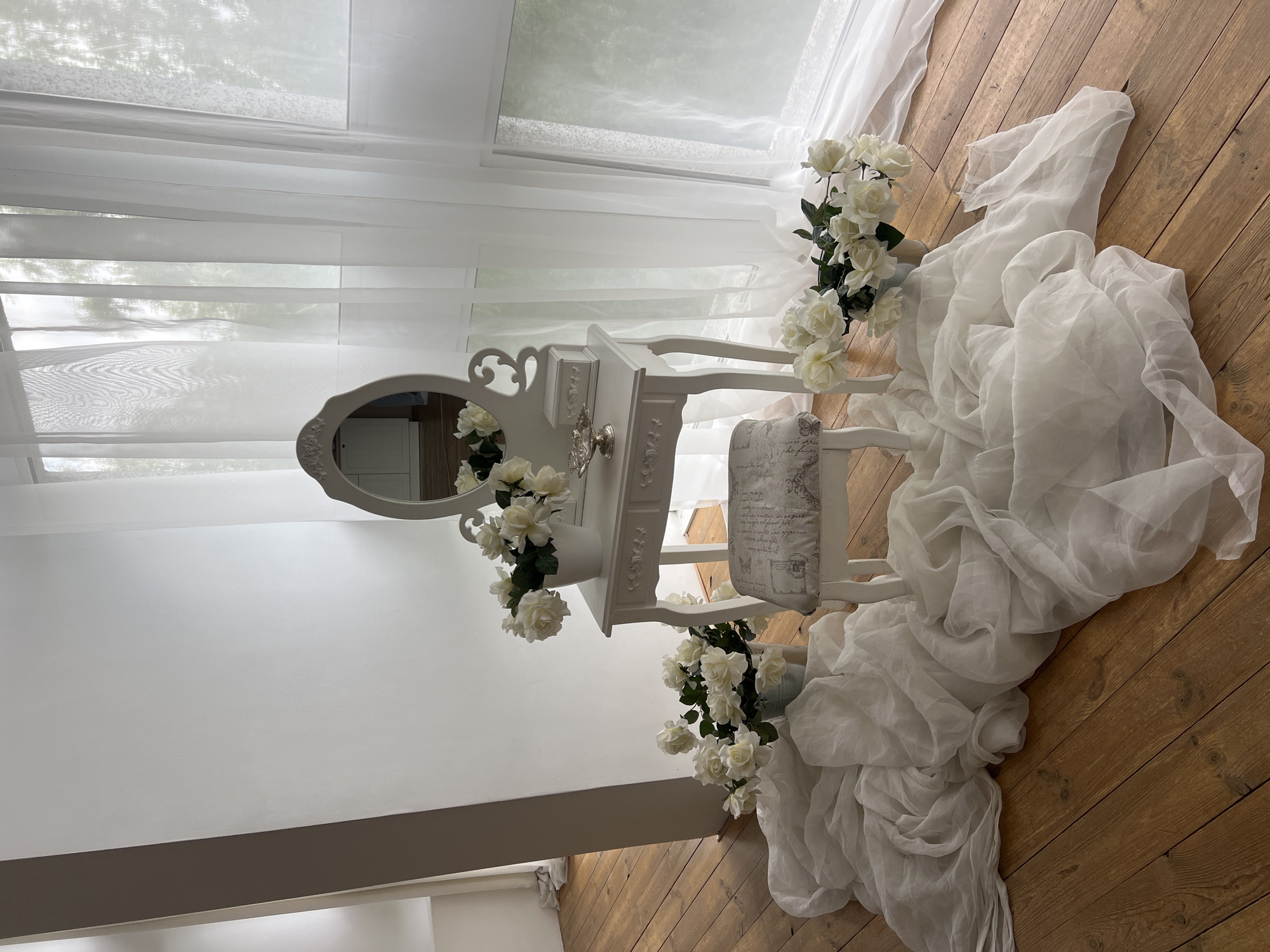 The image depicts a studio setting with a vanity table, stool, and floral arrangements. The vanity table is white with ornate carvings and features a mirror with a decorative frame. On the table, there are two white vases filled with white roses, a small silver jewelry box, and a silver decorative bowl. A stool with a patterned cushion sits in front of the vanity. White sheer fabric is draped around the vanity and stool, pooling on the wooden floor. Two additional buckets of white roses are placed on the floor beside the vanity. The background consists of a plain white wall on the left and large windows covered by white sheer curtains on the right, revealing a blurred view of greenery outside. The floor is made of polished wooden planks. There are no people or animals visible in the image. The scene appears to be indoors, likely in a photo studio, and there is no specific information within the image to suggest a location in Kyiv, Ukraine.