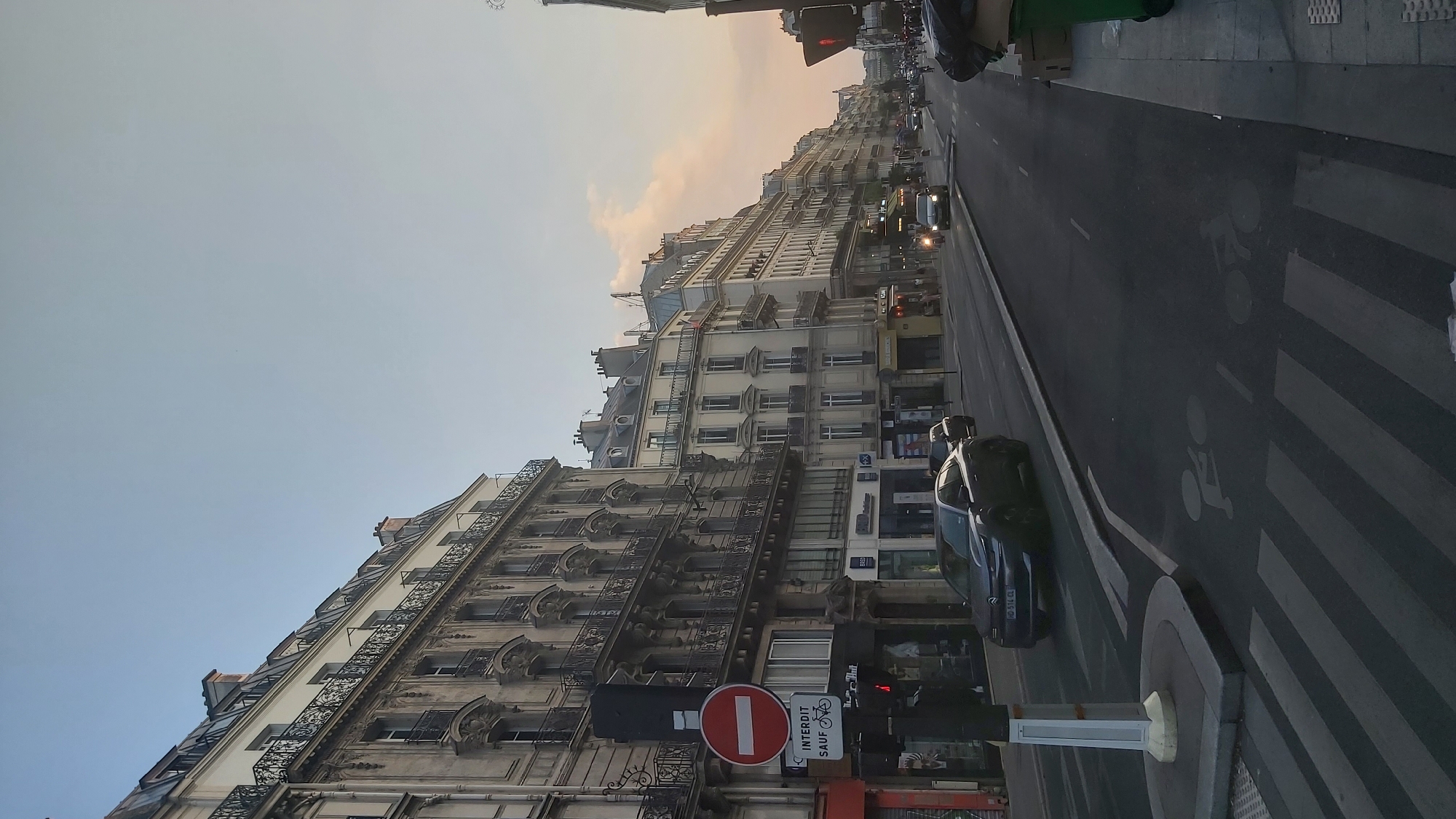 This media file captures a street scene in Paris, France, likely during the late afternoon or early evening. The sky is a soft gradient of pale blue to warm peach, suggesting a sunset or sunrise. The perspective is from street level, looking down a Parisian boulevard lined with classic Haussmann-style buildings. These buildings are characterized by their cream-colored facades, mansard roofs, ornate balconies with wrought-iron railings, and numerous windows.
The street is bustling with traffic, including cars and what appears to be a bus or larger vehicle. Pedestrians can be seen on the sidewalks and crossing a pedestrian zone marked with white stripes. A prominent traffic light with a red signal is visible, indicating a pause in the flow of vehicles. Near the traffic light, a circular red sign with a white horizontal bar, signifying "no entry" or a restriction, is displayed alongside a sign indicating "INTERDIT SAUF BICYCLETTE" which translates to "FORBIDDEN EXCEPT FOR BICYCLES." The road also features markings for a bicycle lane.
Several cars are parked or moving along the road, including a dark sedan with a license plate that appears to read "MD 574 CL". The scene conveys the everyday activity of a European city, with the architecture hinting at a historic and established urban environment. The overall mood is one of quiet urban movement under a gentle sky.