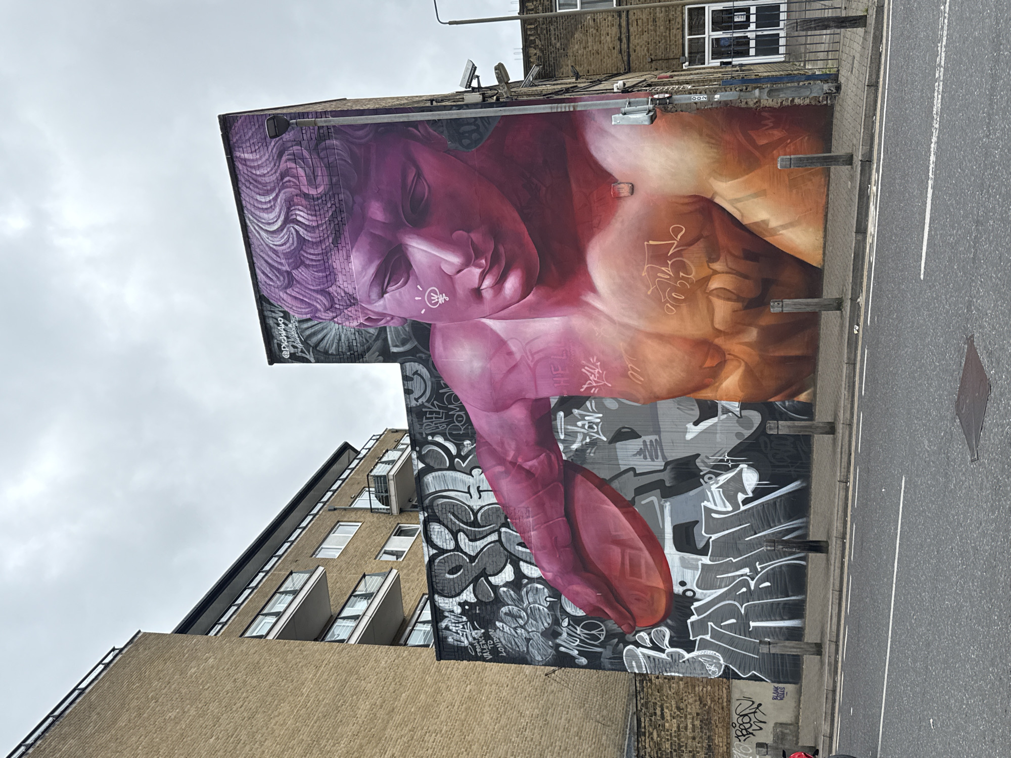 The image depicts a large mural on the side of a building in Shoreditch, London. The mural features a classical figure with purple hair and skin that transitions to orange. There are graffiti elements incorporated into the mural and on the wall below it. The building is brick and has multiple windows. The mural is situated next to a road with a white line marking the edge. Another building, also brick, is visible next to the mural. The sky is overcast.