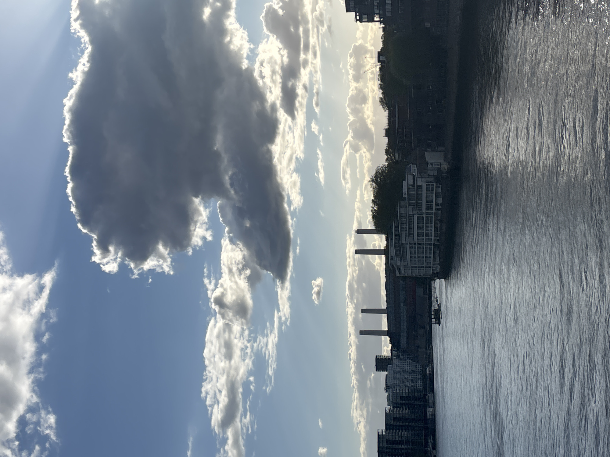 The image shows a view of the River Thames in London, United Kingdom, on a partly cloudy day. The Battersea Power Station is visible with its four chimneys, surrounded by modern buildings. The river is in the foreground and reflects the sky and buildings. A large, dark cloud dominates the left side of the sky.