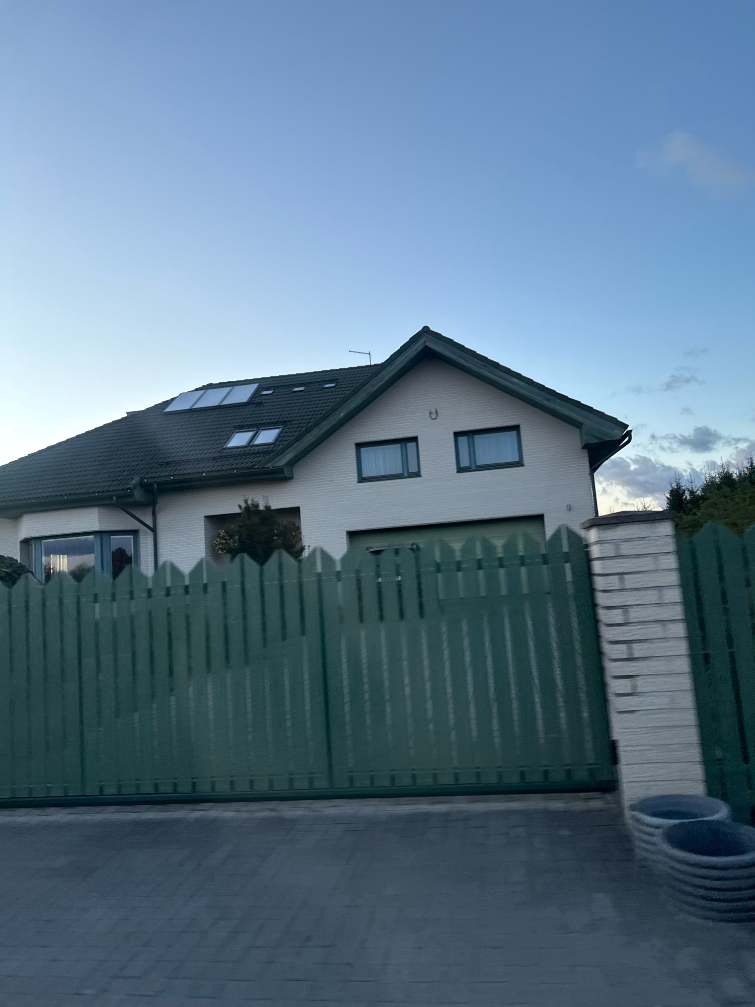A side view of a white, two-story house with a dark green roof is visible in Alliku, Estonia. The house has multiple windows, including several skylights on the roof. A dark green wooden fence runs across the foreground, partially obscuring the house. To the right of the fence, there is a section of white brick pillar and two stacked concrete planters. The sky above is a clear blue with some wispy clouds. The ground in front of the fence is paved with gray interlocking bricks. The overall scene depicts a residential property.