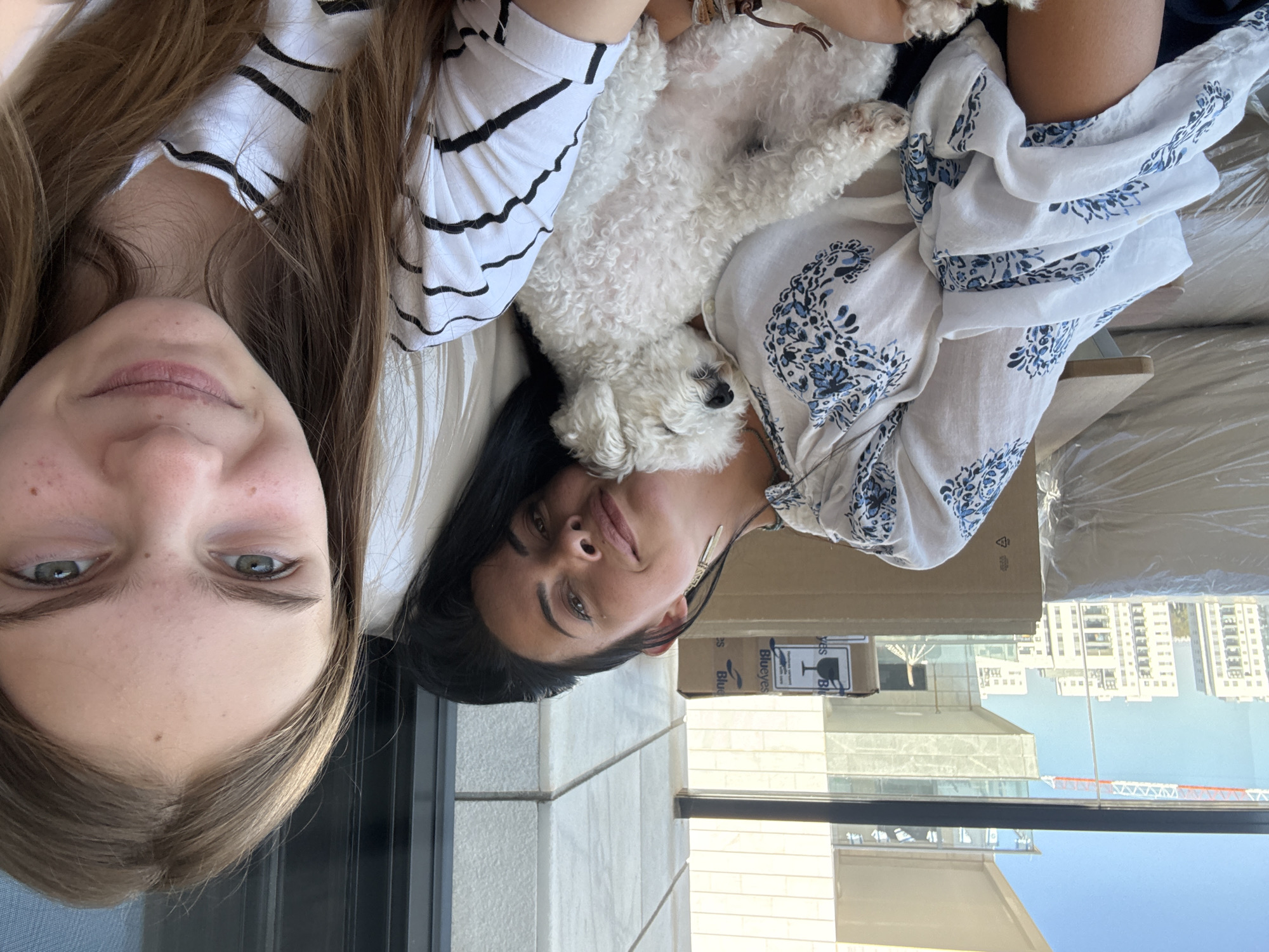 In the city of Or Yehuda, Israel, two females and a small white dog are captured in a selfie. One female, with dark hair, rests her head on a white pillow, a small white dog nestled in her arms, asleep. The other female, with long light brown hair, is in the foreground, her face slightly turned towards the camera. Both are dressed casually. The setting appears to be a balcony or patio, with a cityscape visible in the background, including modern buildings and construction cranes. Cardboard boxes are also present in the scene.