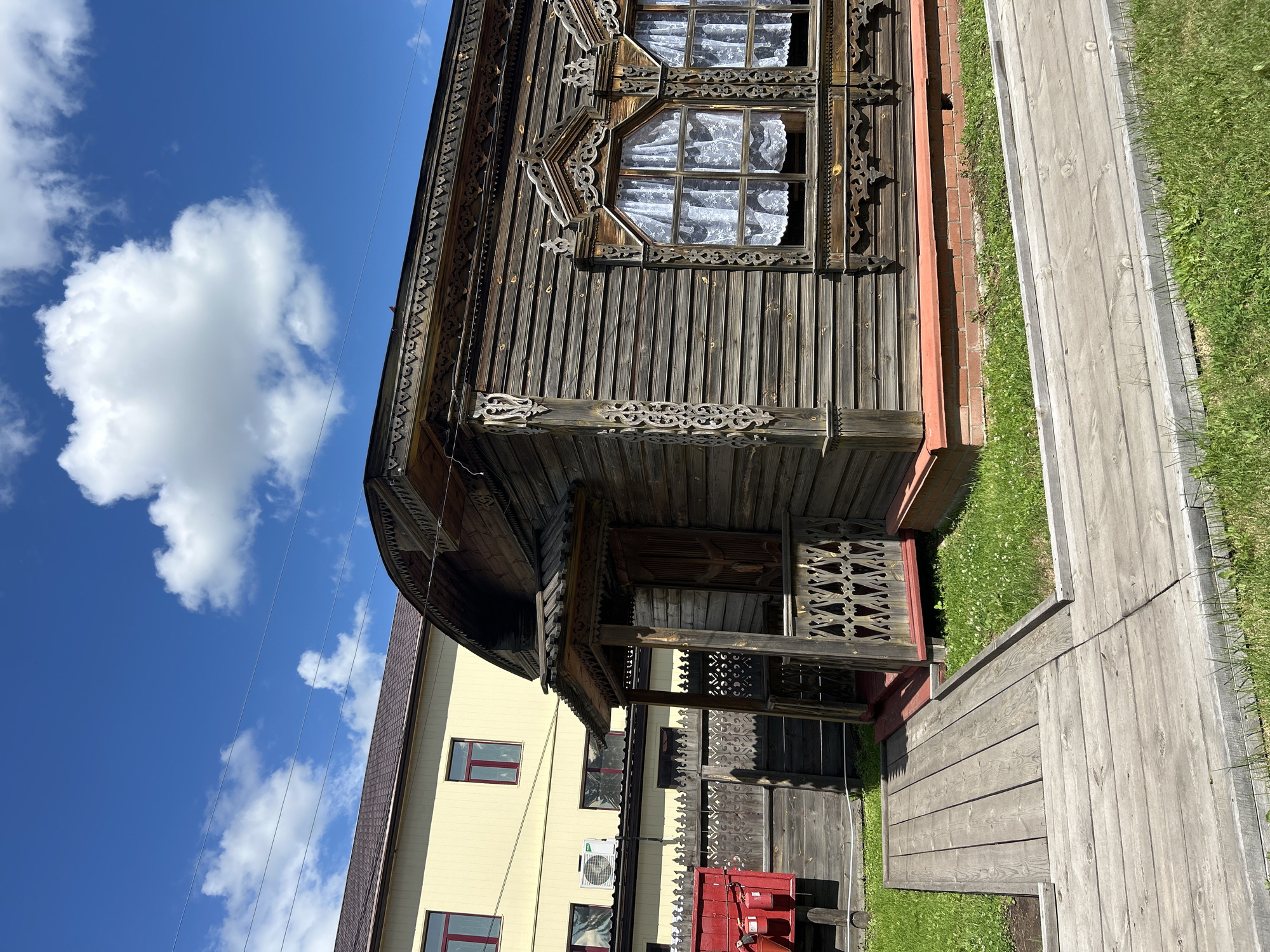 The image depicts the exterior of a historic wooden house in the city of Bolsherechye, Russia. The house features intricate wooden carvings around its windows and eaves, with lace curtains visible through the windows. A small wooden porch with a patterned railing leads to the entrance. In front of the house, a wooden walkway is laid out over a patch of green grass with small white flowers. Behind the wooden house, a modern yellow building with red-trimmed windows is visible, along with a wooden fence and a red metal box, possibly a fire alarm. The sky is a clear blue with scattered white clouds. The scene suggests a warm, sunny day.