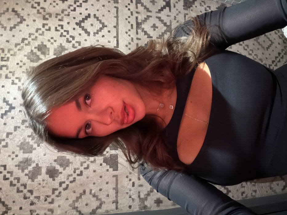 A young woman with shoulder-length brown hair and fair skin is pictured against a textured, patterned wall. She wears a long-sleeved black dress with a chest cutout. She has dark eyes and full lips, and she is looking directly at the camera with a slight pout. A delicate necklace and a thin body chain are visible. The image has warm, slightly reddish lighting.