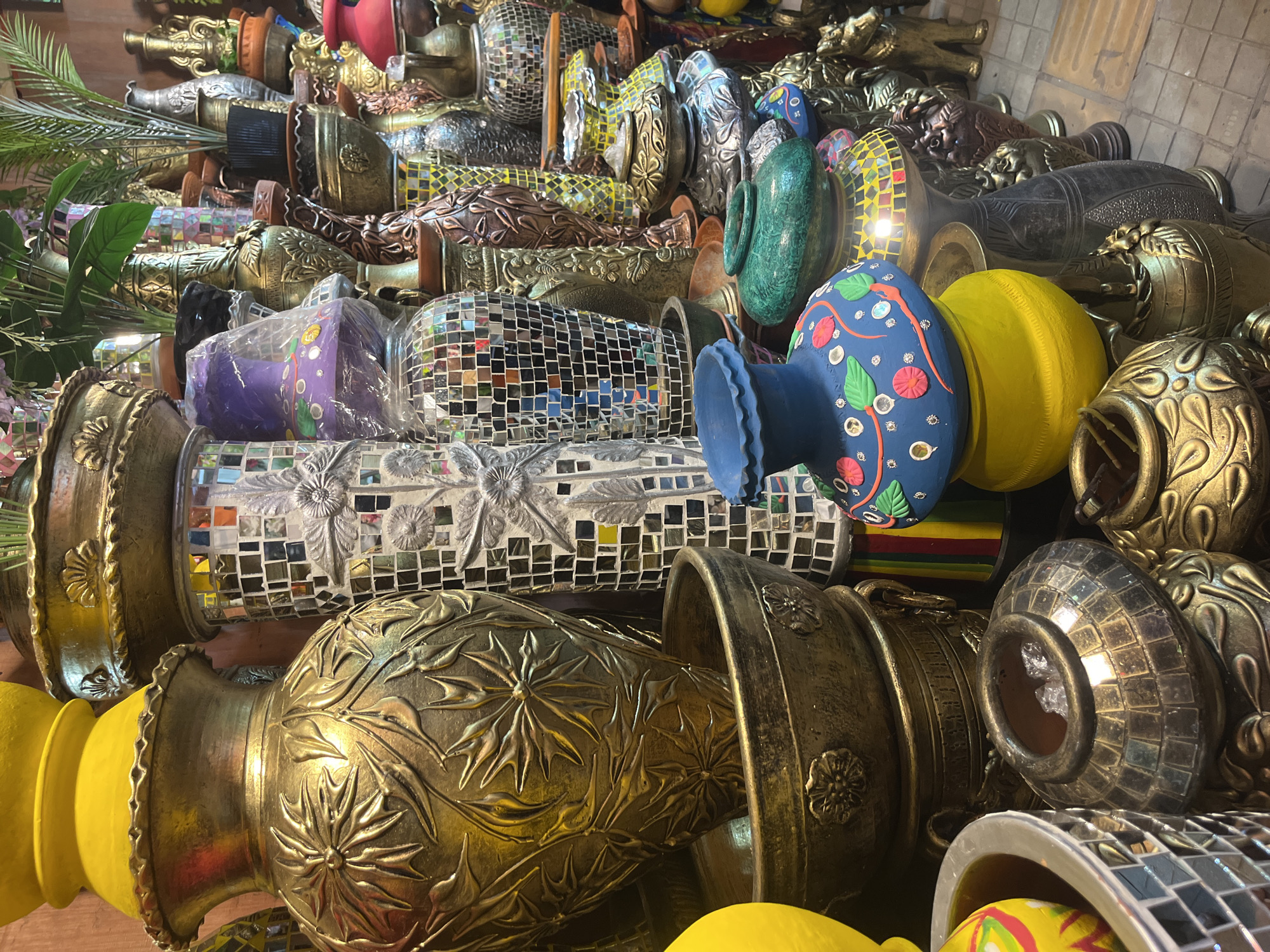 The image shows a large collection of ornamental vases and pots of various sizes, shapes, and materials. These are primarily made of metal and ceramic, with mosaic details. Some are painted in bright colors like blue and yellow, while others have metallic finishes such as gold and bronze. They appear to be arranged in a cluttered, stacked manner, suggesting a retail setting or storage area. The scene could be a shop or market stall in Dhaka, Bangladesh, known for its vibrant marketplaces and crafts.