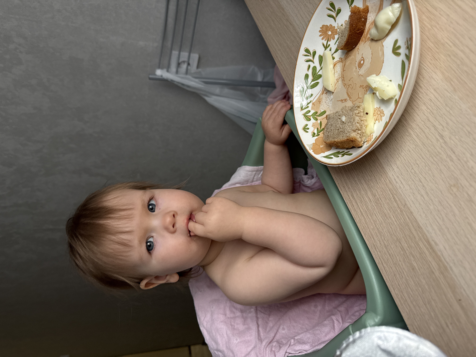 A baby sits in a high chair in what appears to be an indoor setting. The baby is fair-skinned with light brown hair and blue eyes, and is shown from the waist up, unclothed except for a light pink cloth draped around the lower body. The baby is looking towards the camera with its right hand near its mouth. To the right of the baby is a wooden table with a decorative plate. On the plate, there are pieces of bread and cheese. Behind the baby, there is a grey wall. The location of the city of Moscow, Russia, is not discernible from the image.
