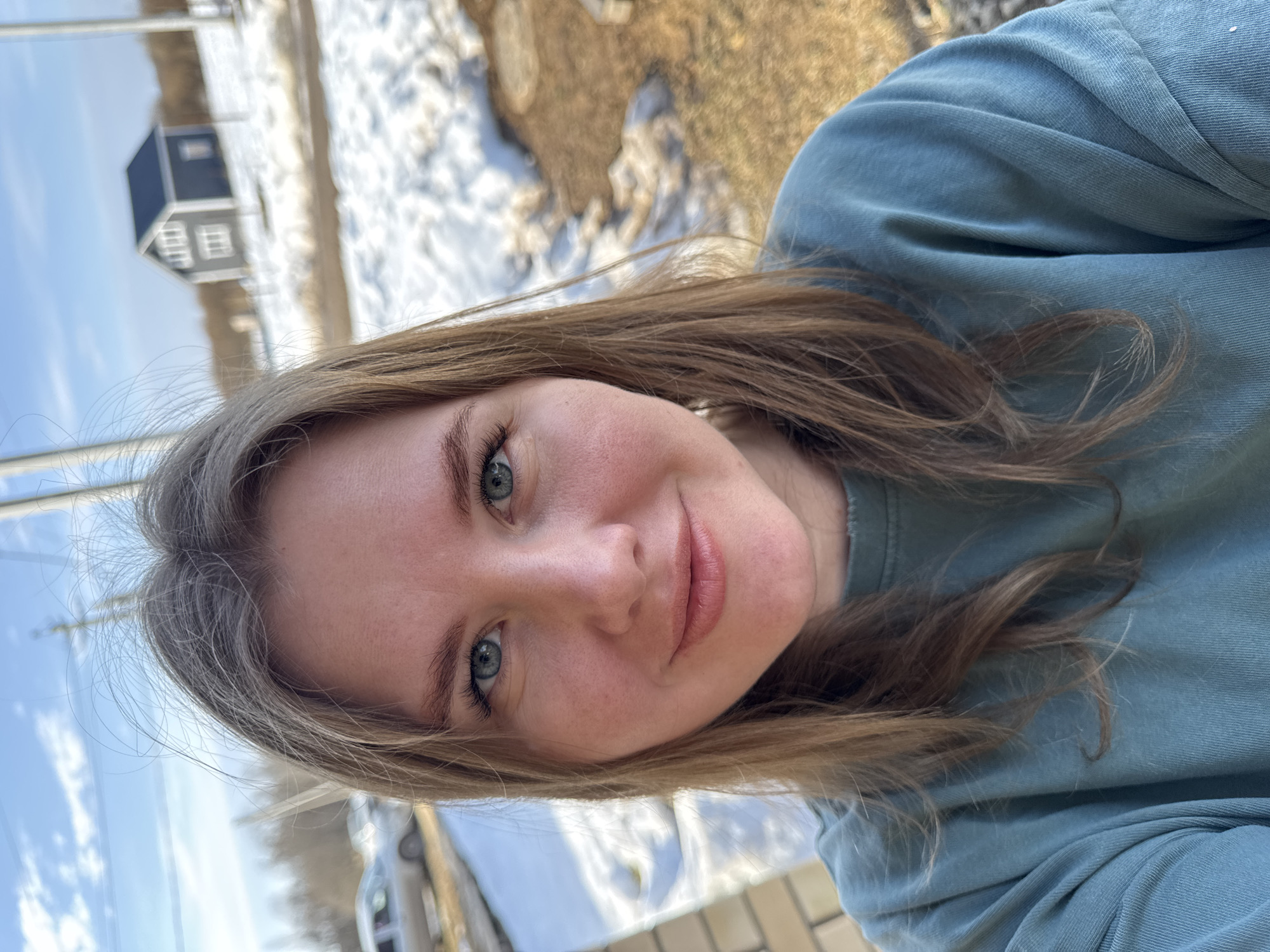 A woman with light brown hair and blue eyes is posing for a selfie in Gornyi (Dvurechenskoe S/P), Russia. She is wearing a blue-green top. In the background, there is a wooden house with a dark roof and white windows, snow, and grass. A vehicle is also visible in the background.