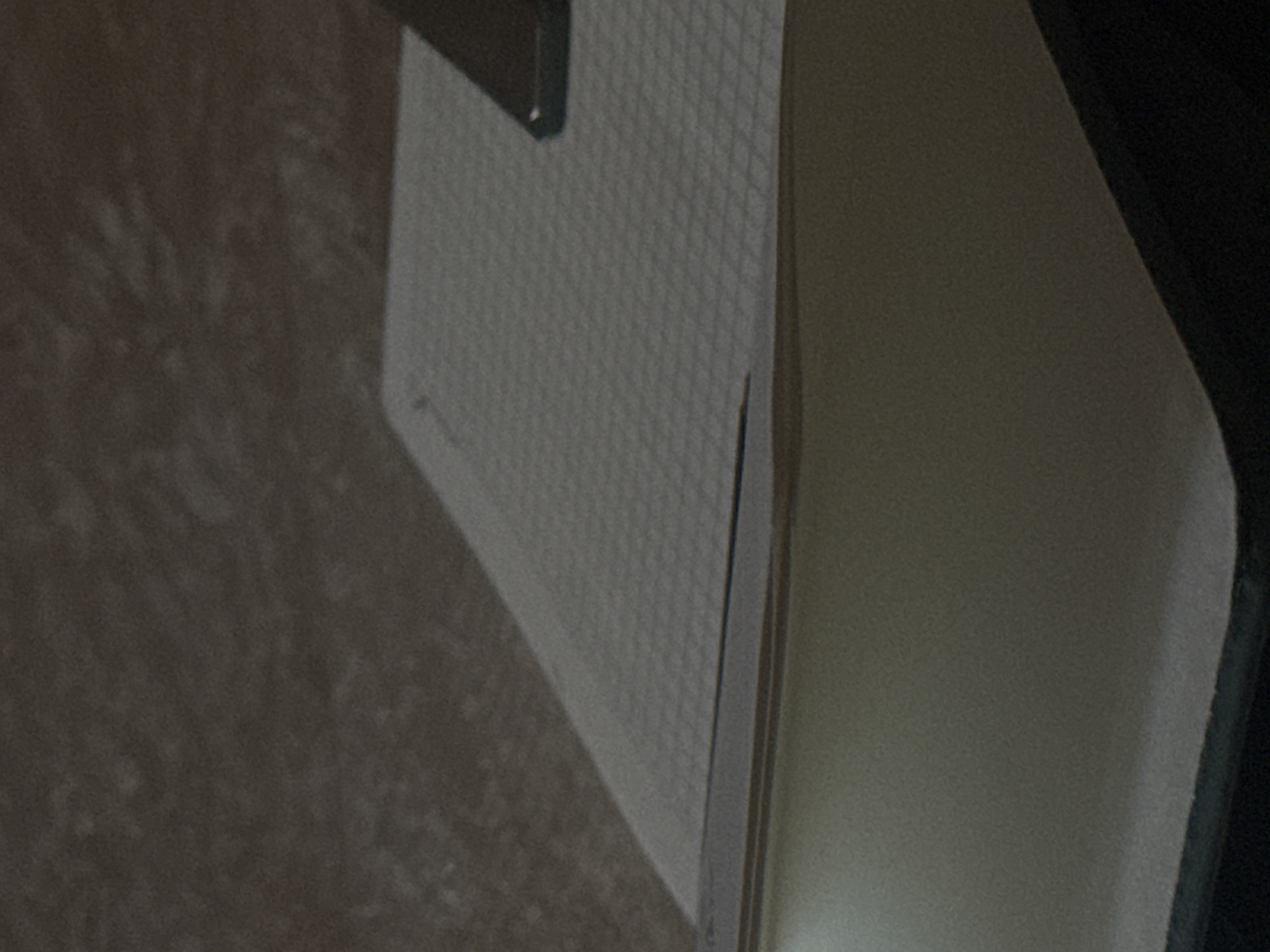 A close-up shot shows a white surface with a grid pattern, possibly a notebook or a pad of paper. A dark object, which appears to be a smartphone, is resting on the upper right corner of the white surface. The background is a textured, dark gray surface. The scene is likely indoors, with soft lighting that creates subtle shadows. No people are visible in the image. The location of Navbahor, Uzbekistan, is not discernible from the visual information provided in the image.
