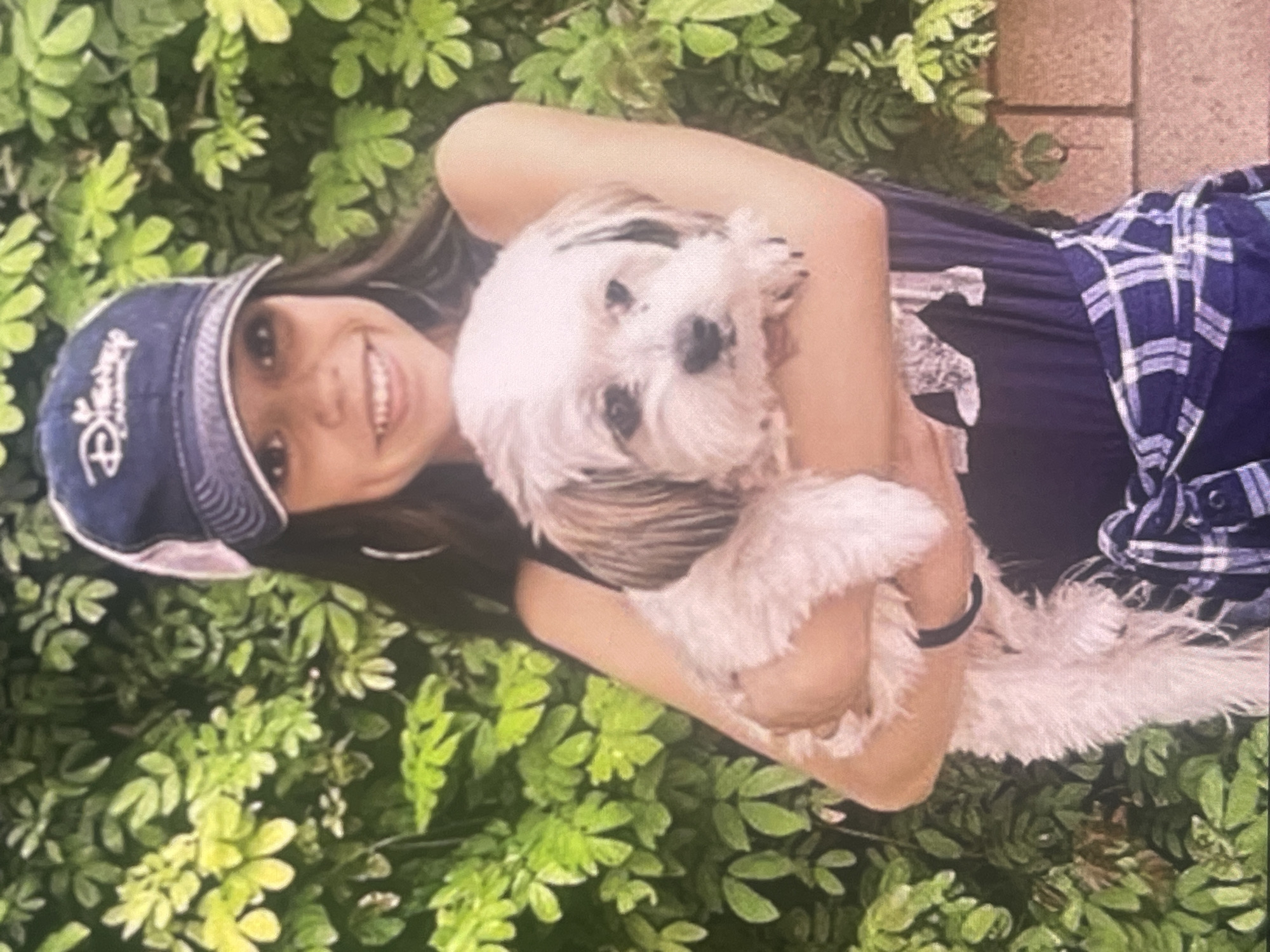 A young person with dark hair and a wide, joyful smile holds a small, fluffy white and brown dog outdoors in Poltava, Ukraine. The person is wearing a dark blue t-shirt and a dark blue baseball cap with white, stylized text that appears to read "Disney." A blue and white plaid shirt is tied around their waist or arm. They have a black bracelet on their right wrist. The dog, likely a Shih Tzu or similar breed, has dark markings around its eyes and nose and is looking towards the camera with a neutral expression. The background consists of vibrant green foliage, suggesting a garden or park setting, with a visible brick or paved surface on the right side. The scene is brightly lit, indicating it is daytime. The overall mood is cheerful, reflecting the person's happy expression.