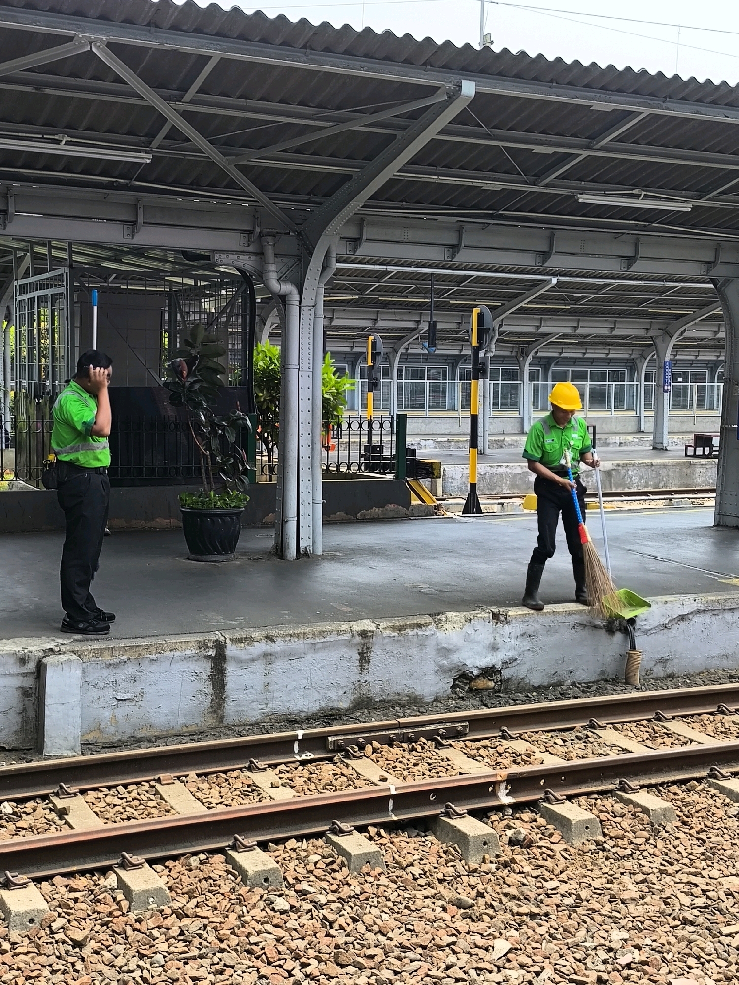 Two male workers are present at a train station. On the left, a worker in a green shirt and black pants stands with his hand on his head, looking downwards. On the right, another worker wearing a yellow hard hat, green shirt, and black pants is actively sweeping debris from a platform onto a dustpan with a broom. The environment appears to be an outdoor covered train platform with overhead lighting and structural supports. In the foreground, train tracks are visible, filled with ballast stones. The station setting suggests an urban location, likely in West Jakarta, Indonesia, given the context.