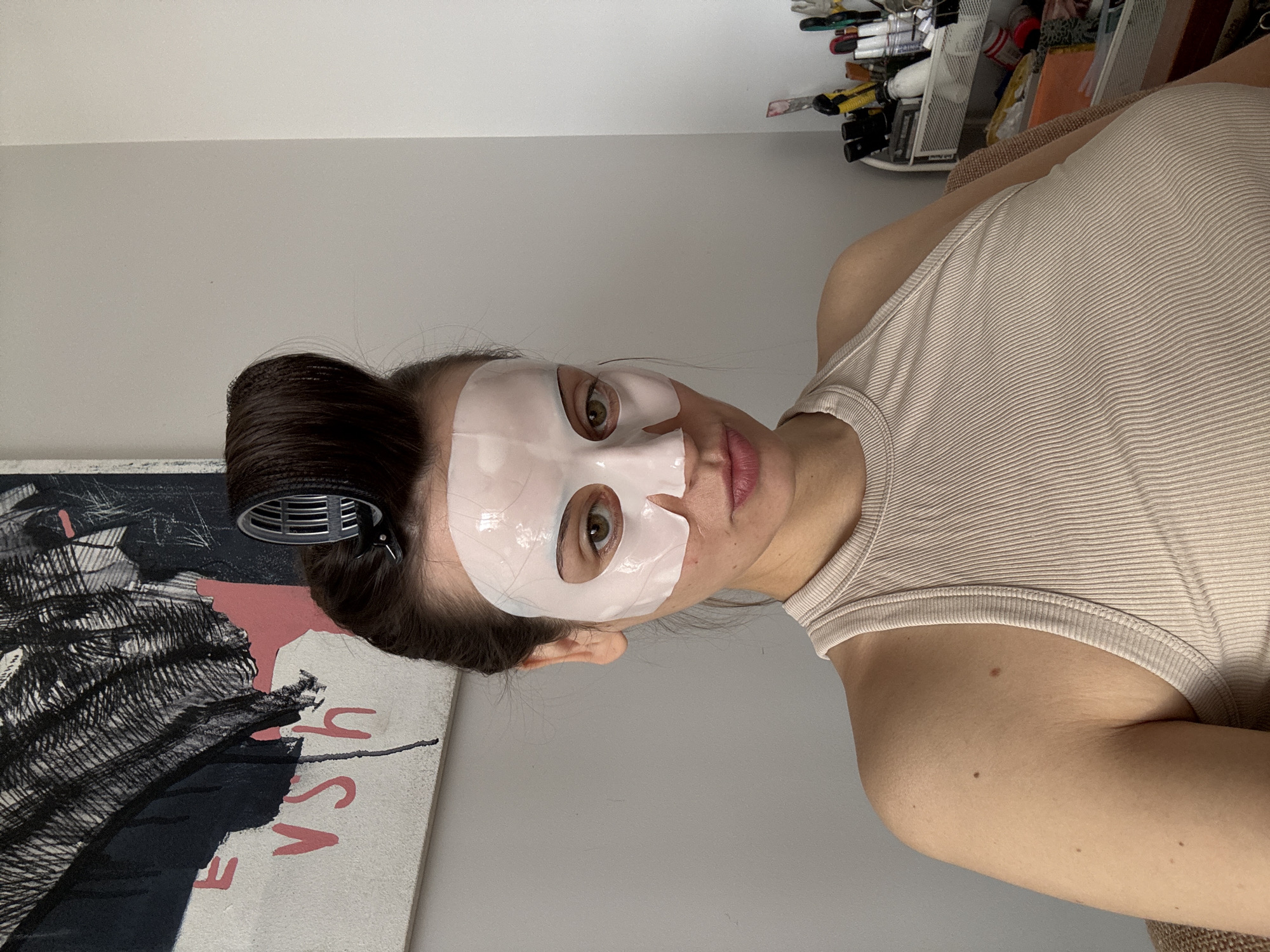 A woman is lying down wearing a sheet face mask and a beige tank top. Her hair is in a bun with a hair clip. There is artwork on the wall behind her, and a container with art supplies is visible in the background. The location is unknown, and there is no indication of it being Krasnoyarsk, Russia.