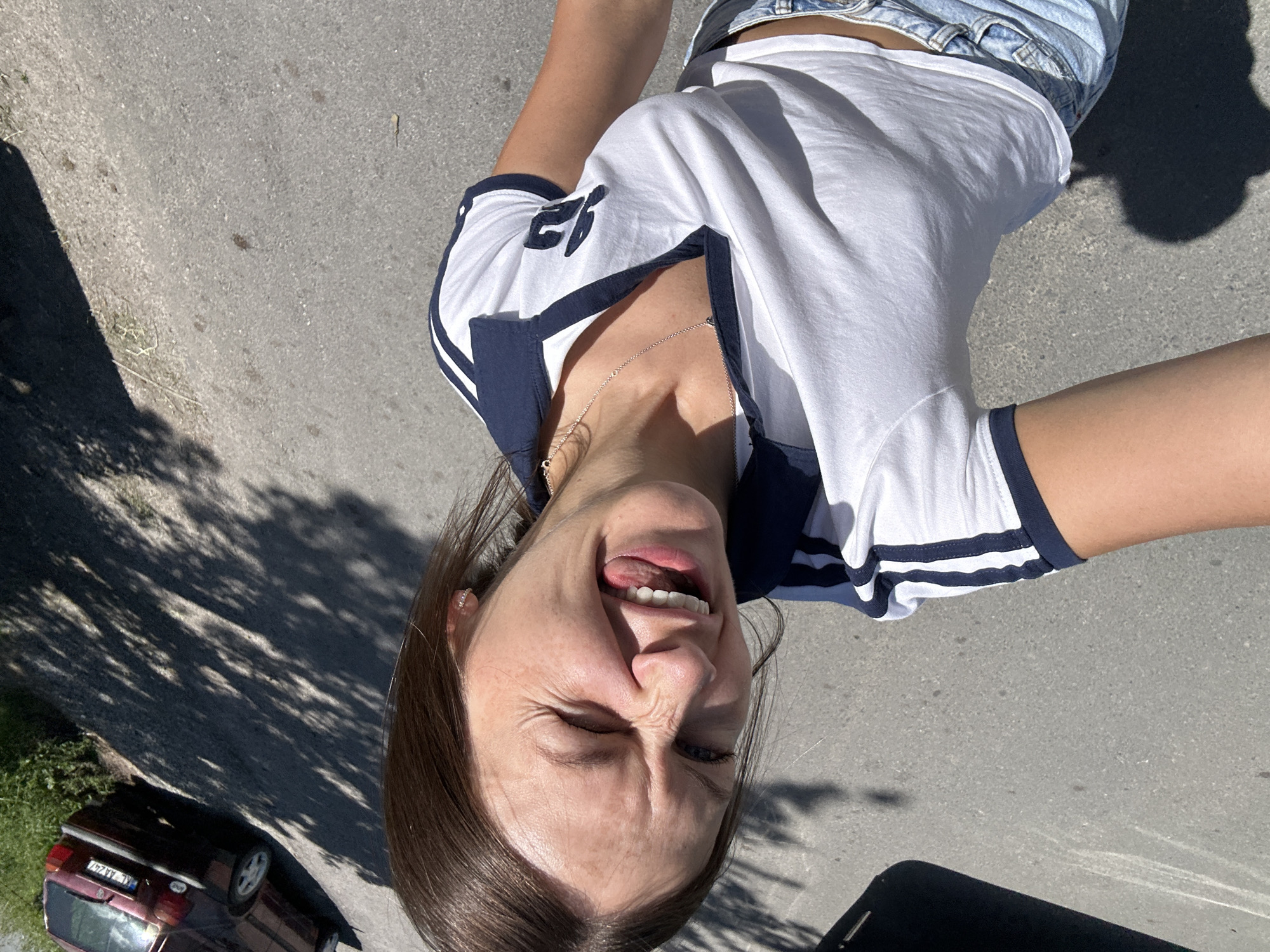A young woman is photographed from a high angle, outdoors, with a road and shadows visible. She is wearing a white t-shirt with navy blue trim and blue denim shorts. Her eyes are closed, and her tongue is sticking out as she smiles. A maroon car is parked on the left side of the frame, and green foliage is visible in the background. The scene appears to be in the city of Kolašin, Montenegro.