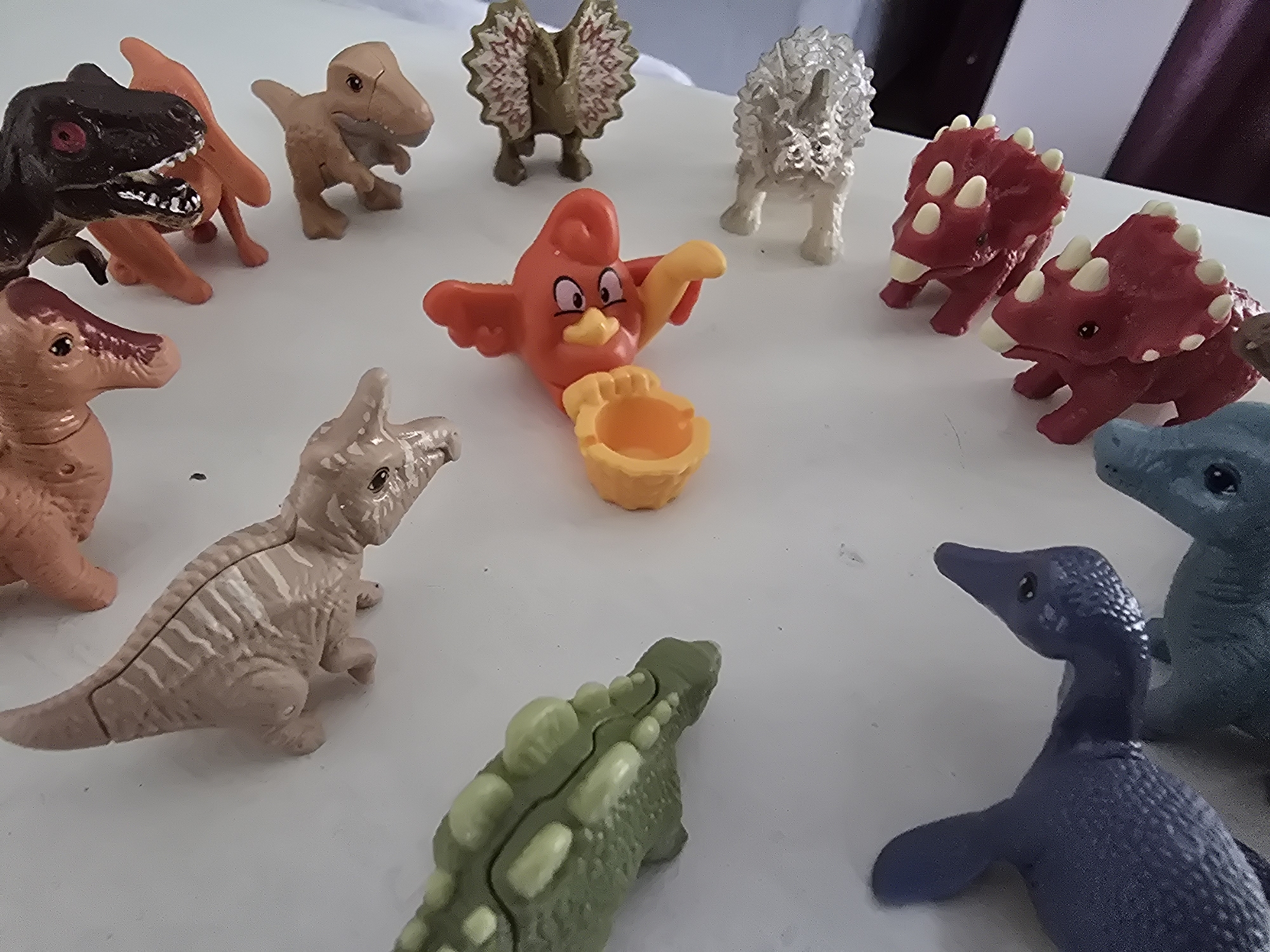 The image shows a collection of plastic dinosaur figurines arranged on a white surface. There are several different dinosaur species represented, including a Tyrannosaurus Rex, Triceratops, Dilophosaurus, and Plesiosaur. Additionally, there is a cartoon bird figure holding a yellow cup-like object. There are no people present. The location is not discernible from the image.