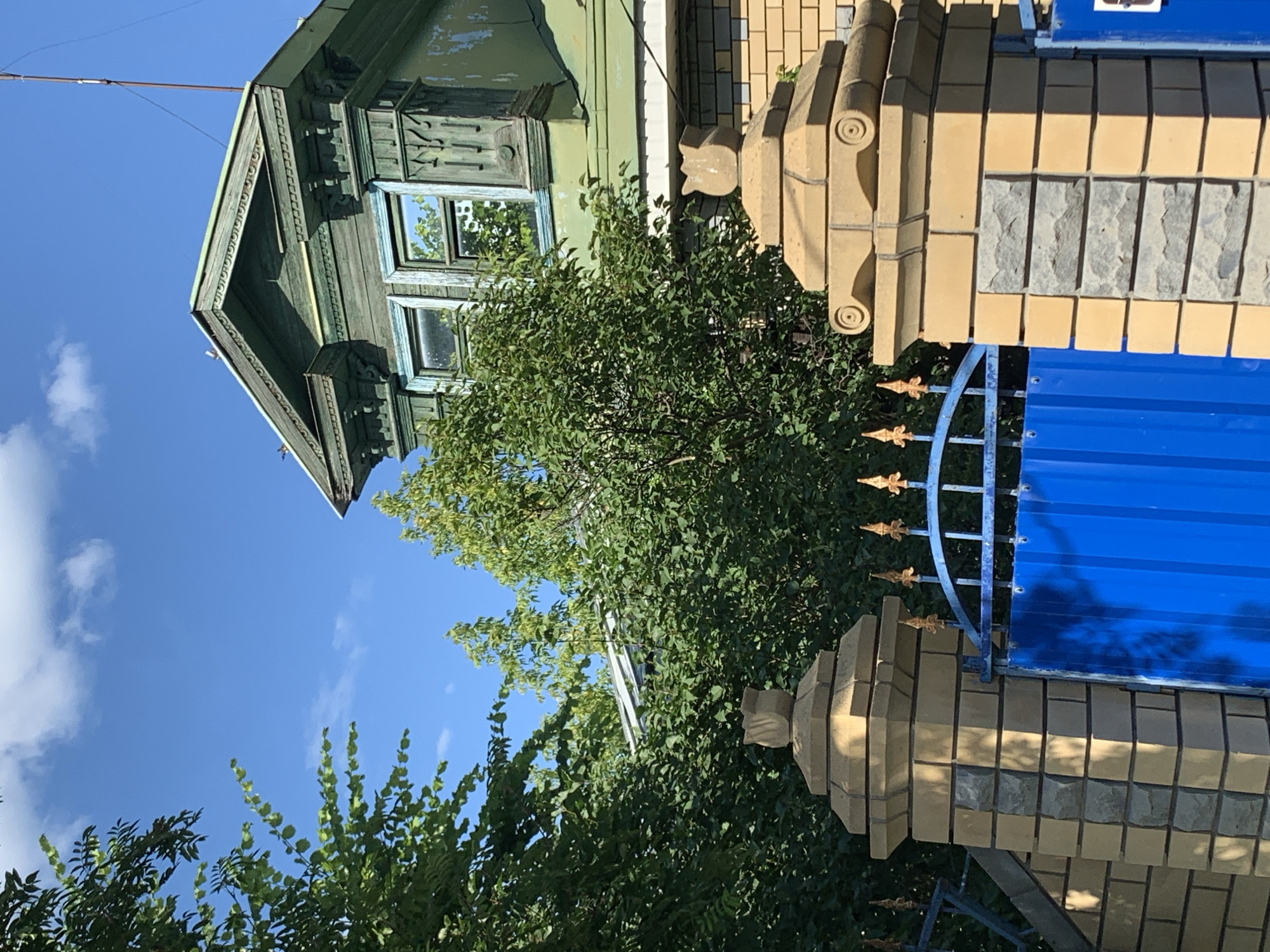 The image captures an exterior view of a traditional Russian wooden house, painted green and showing signs of age, with ornate trim around the windows and eaves. The house is situated next to a fence made of light-colored bricks and topped with a bright blue metal sheet. Decorative elements are visible on the brick pillars and a golden metal gate. Lush green trees and foliage surround the property, suggesting a natural or garden setting. The sky is a clear, bright blue with a few scattered white clouds, indicating a sunny day, likely during daylight hours. The composition and subject matter suggest a residential setting, possibly in a rural or suburban area of Pavlovo, Russia. There is no visible text in the image.