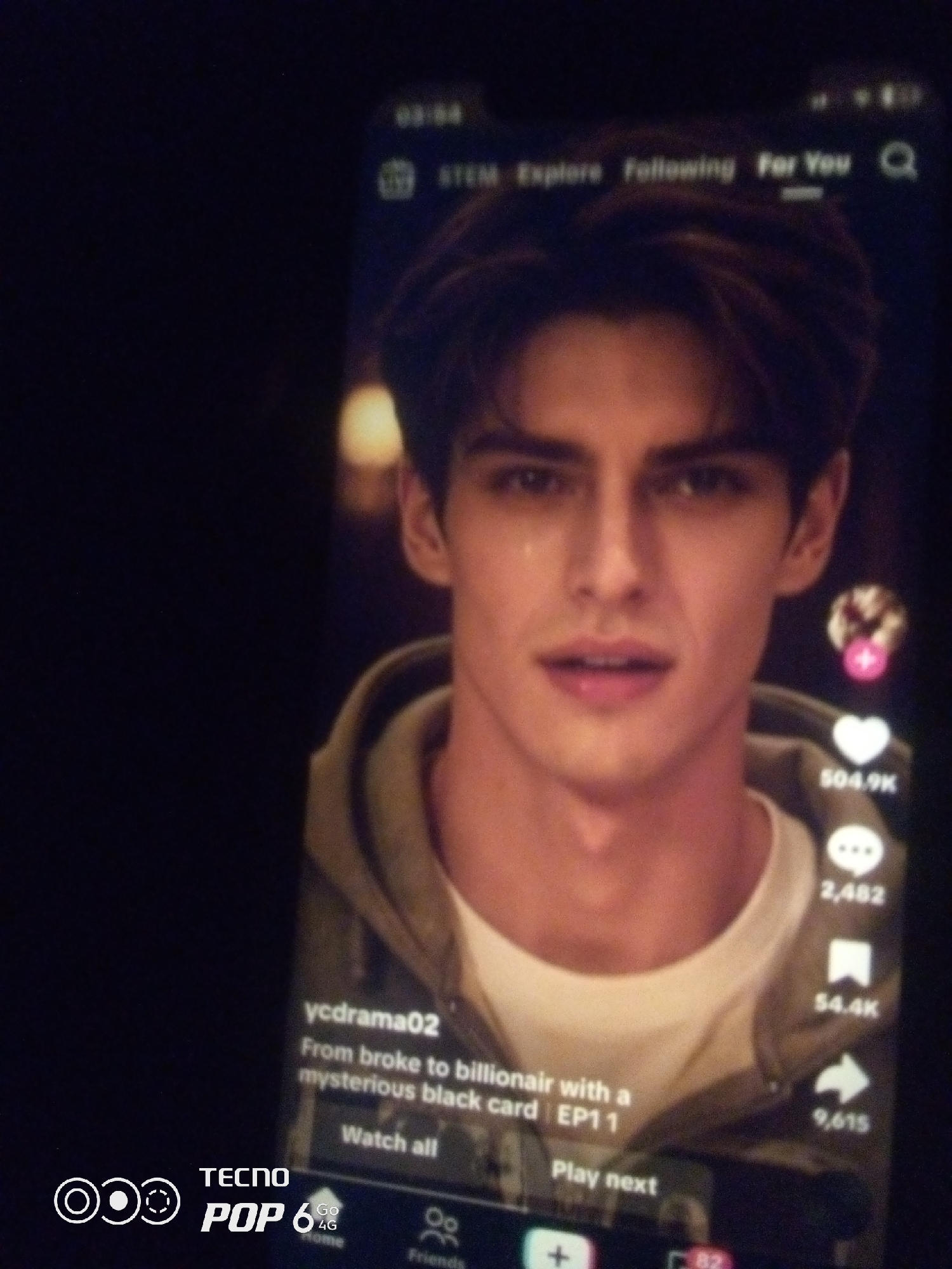 The image shows a close-up of a smartphone screen displaying a social media video. The video features a young man with dark hair and a light-colored hoodie. Text overlays on the video indicate a username "ycdrama02" and a caption "From broke to billionair with a mysterious black card | EP1 1". Below this, there are buttons for "Watch all" and "Play next". On the right side of the screen, there are icons for likes (heart icon with 504.9K), comments (speech bubble icon with 2,482), shares (arrow icon with 9,615), and a bookmark icon (with 54.4K). The top of the screen shows navigation options including "STEM", "Explore", "Following", and "For You". The bottom of the screen displays a navigation bar with icons for home, friends, and other unspecified actions, along with a "TECNO POP 6 4G" logo. The image itself appears to be a photograph of the phone screen taken in a dimly lit environment, with a dark, out-of-focus background. There is no visible indication of the city of Howayi, Nigeria within the image content.