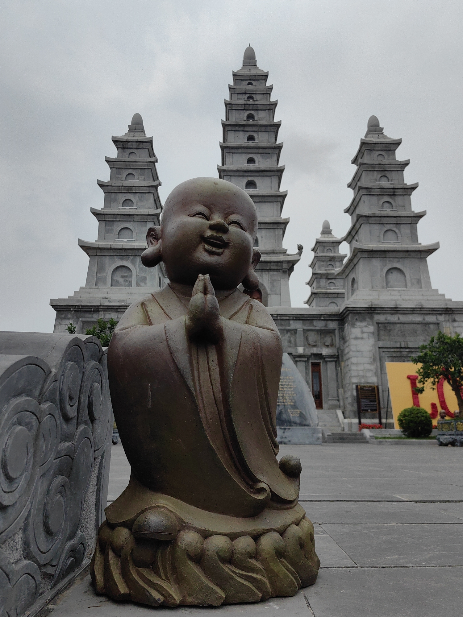 A smiling, brown, child-like statue is the focus of a ground-level view. Its hands are pressed together in a prayer-like gesture. The statue is situated next to a gray stone railing. Behind the statue, three tall, gray, pagoda-style structures rise against an overcast sky. The background reveals a building with steps and decorative elements. A glimpse of a yellow sign and foliage is also visible. The scene is set in Hong Gai Ward, Vietnam. The location is outdoor and the weather is cloudy.