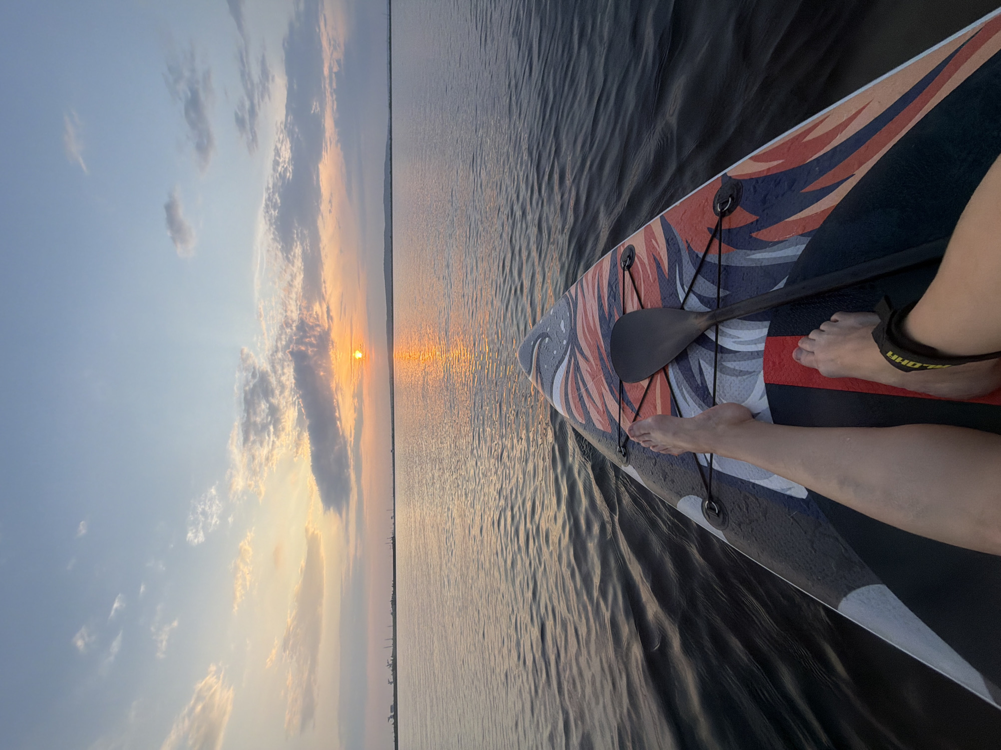 The image depicts a first-person perspective of someone on a paddleboard on a body of water, likely the Kama River, near Chaikovsky, Russia, during sunset. The sky is a blend of pale blue, orange, and pink hues, with scattered clouds. The sun, a bright orange orb, is visible low on the horizon, casting a golden reflection on the rippling water. In the distance, a line of industrial structures and trees is silhouetted against the sky. The foreground is dominated by the paddleboard, which has a colorful, abstract design. A paddle lies on the board, and a person's legs and feet are visible, with one foot bare and the other wearing a black ankle strap. The water is dark and calm, with gentle waves.