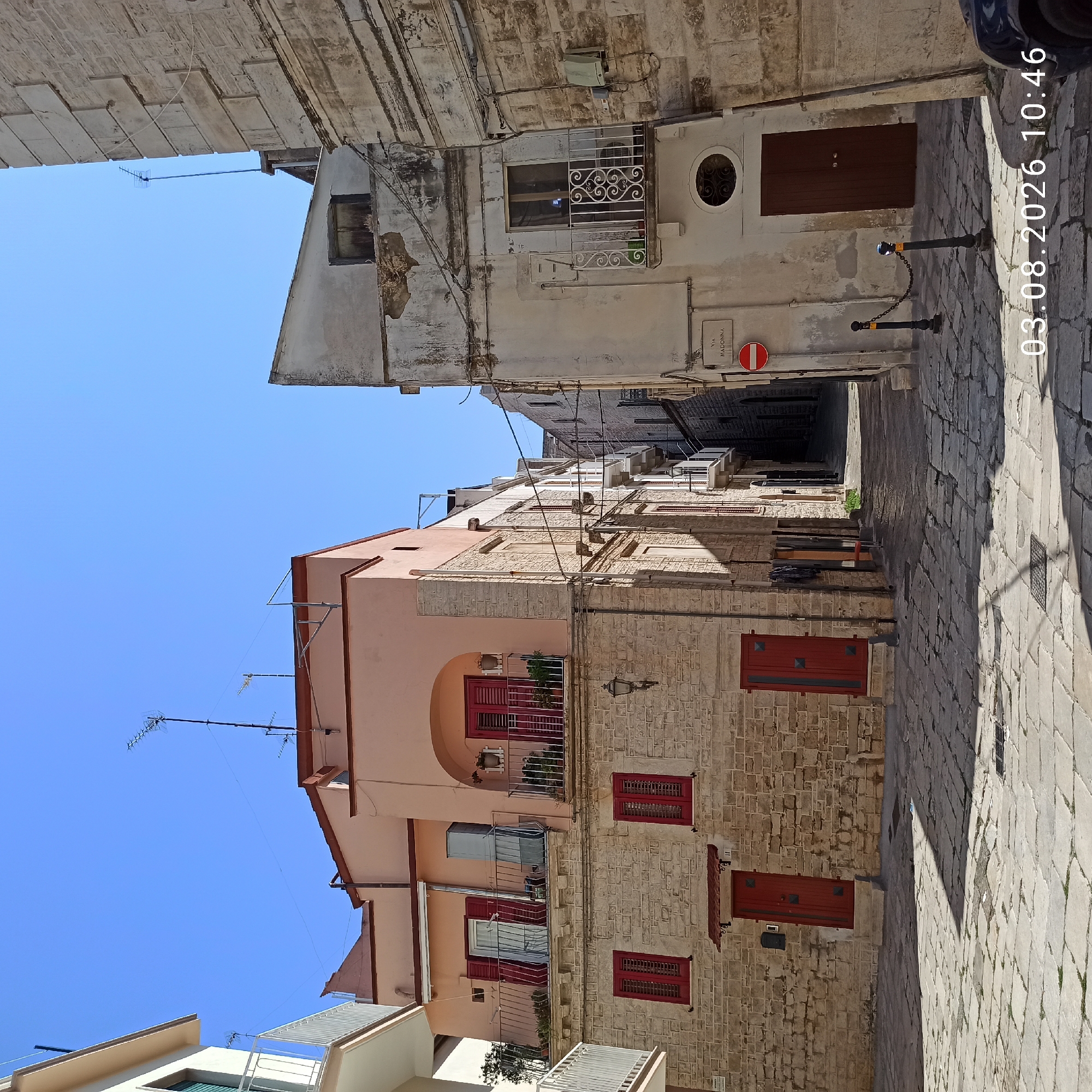 The image depicts a sunlit street scene in the historic center of Ruvo di Puglia, Italy, likely near Piazza Dante. The architecture is characteristic of the region, with stone buildings and light-colored stucco facades.
On the left, a building with a pale pink upper story and a stone lower story features red-shuttered windows and a balcony with red railings. Two red doors are visible on the ground floor. To the left of this building, a portion of a modern white building with a balcony is visible.
Adjacent to the pink building, on the right, stands a weathered, multi-story building with a pale, textured facade. This building has a small balcony with ornate white railings and a dark wooden door. Further down the narrow street, other buildings with balconies and stone facades are visible, receding into shadow.
The street is paved with irregular stone slabs, with visible cobblestones and drainage grates. Several dark metal bollards connected by chains are positioned along the sidewalk. A red and white circular street sign, indicating "VIA MADONNA," is affixed to a pole on the right side of the street. The sky is clear and blue. The timestamp "03.08.2026 10:46" is overlaid on the lower right corner of the image. No people or animals are present. The overall impression is one of a quiet, historic Italian town on a bright day.