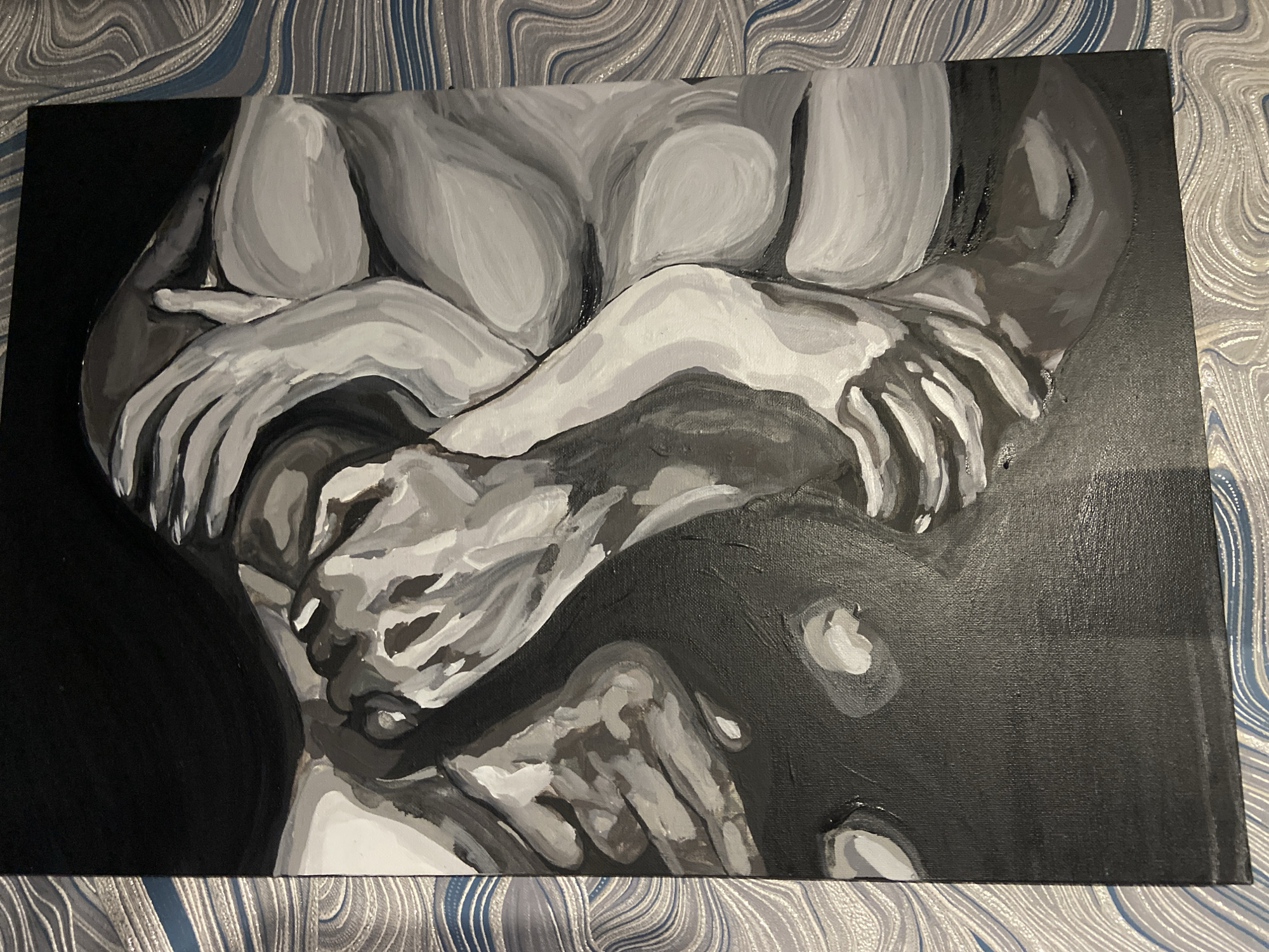 The image is a painting on canvas featuring a black and white abstract depiction of intertwined human forms. The composition is dominated by shades of gray, black, and white, creating a chiaroscuro effect. The forms suggest a close embrace or a single figure folded upon itself, with emphasis on the curves of bodies and the interplay of light and shadow on the skin. Hands are a prominent element, rendered with flowing lines that convey a sense of movement and connection. The background is solid black, which further accentuates the figures in the foreground. The painting is placed on a surface with a marbled pattern of blue and gray. There is no identifiable location in the city of Stavropol, Russia depicted in this image; it is a studio or indoor setting.