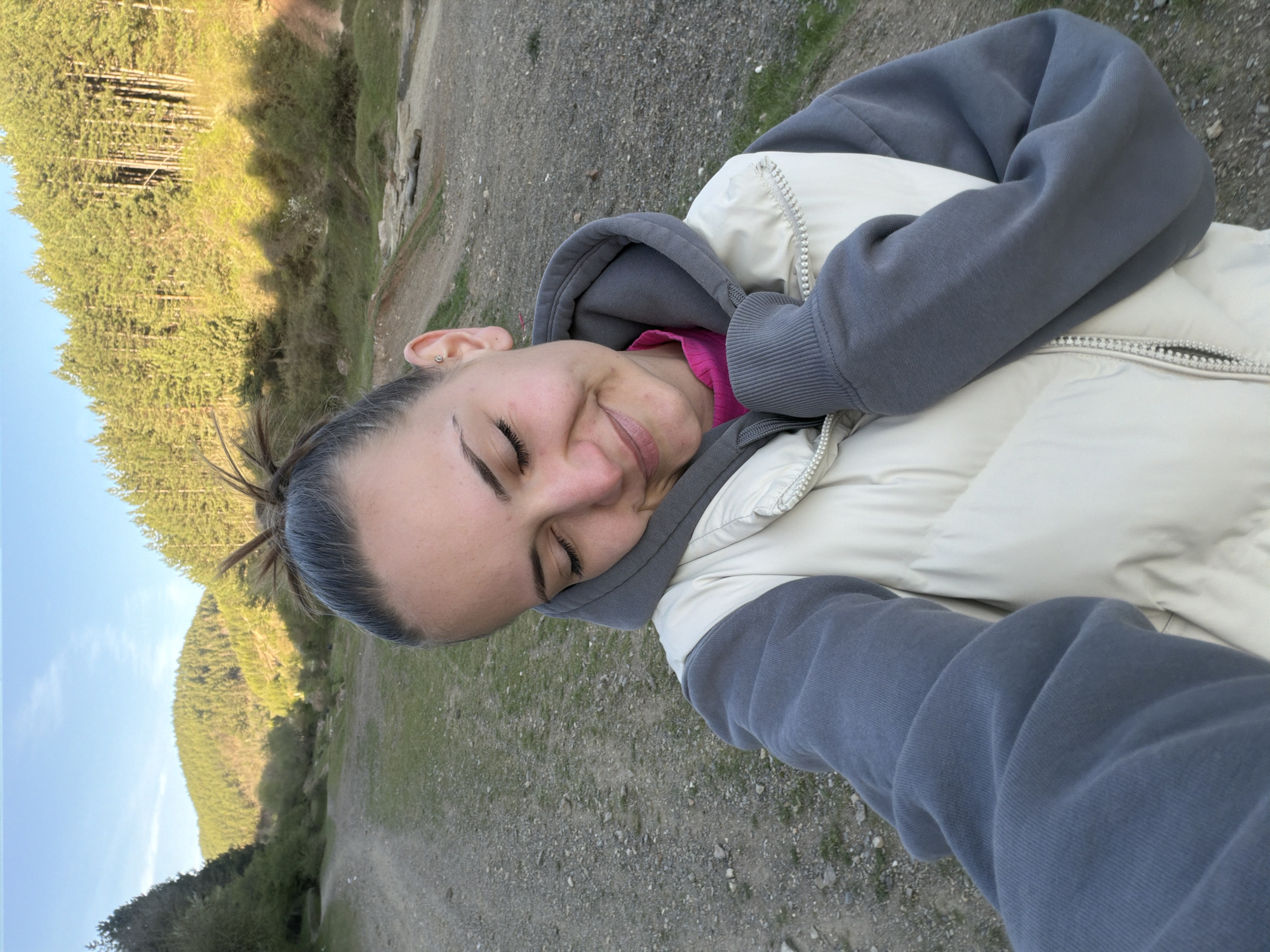The image displays a close-up selfie of a young woman enjoying an outdoor setting in Buhovo, Bulgaria. The woman is positioned on the right side of the frame, smiling with her eyes closed, conveying a sense of peace or contentment. Her hair is pulled back, and she has a small, circular earring in her visible left ear.
She is dressed in warm, layered clothing suitable for an outdoor activity. She wears a grey hooded sweatshirt with the hood up, covering a bright pink top. Over the sweatshirt, she has a light beige or cream puffer vest with visible zippers.
The background reveals a natural landscape dominated by dense evergreen forests covering steep hills or mountainsides. A dirt or gravel path or road runs through the lower part of the scene. The sky, visible in the upper left, is clear and light blue, indicating fair weather. The lighting suggests either early morning or late afternoon, with the sun casting soft light and some shadows on the trees.
The overall scene depicts a person enjoying a moment of tranquility amidst nature, likely during a hike or walk in a mountainous region. There is no visible text in the image.