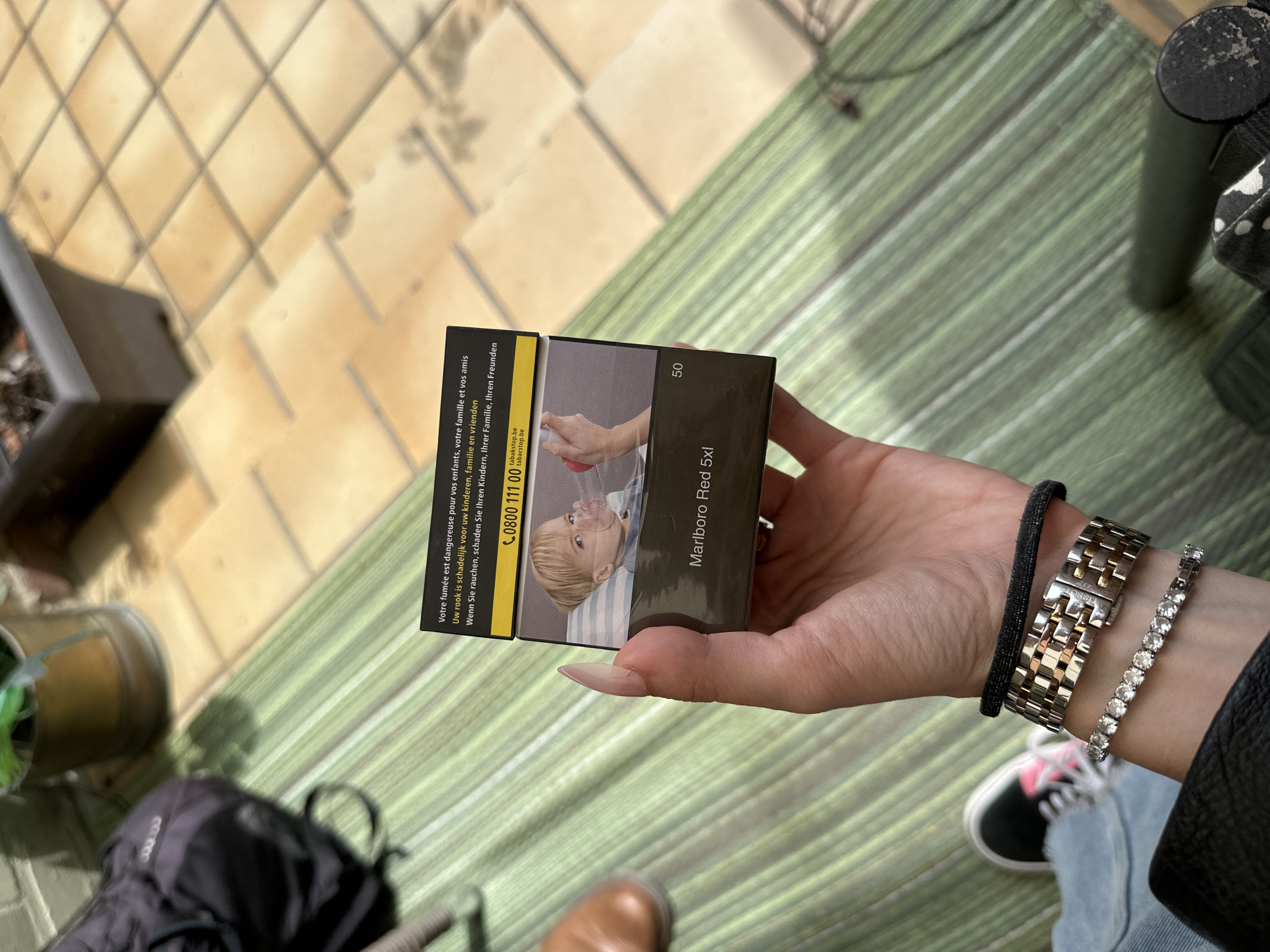 A person's hand, with long, light-colored fingernails, holds a pack of Marlboro Red 5xl cigarettes. The pack features a warning image of a child wearing a breathing mask. The background shows a green striped mat on a tiled patio, with a blurred backpack and a planter in the upper left corner. The lighting suggests an outdoor setting. The image does not provide any information about the city of Bruges, Belgium.