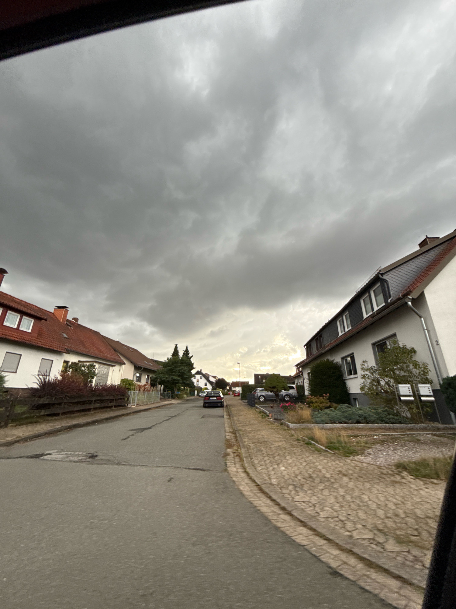 The image shows a residential street in Bad Sachsa, Germany, under a heavy, dark cloud cover. The perspective is from a moving vehicle, looking forward and slightly to the right. On the left side of the street, there are several houses with red tile roofs and white walls. A dark sedan is parked on the right side of the street further down. The right side of the street features a paved walkway made of light-colored cobblestones, and more houses with white walls and dark roofs. The sky is overcast with thick, grey clouds, but a brighter patch of sky is visible in the distance towards the horizon, suggesting the sun may be trying to break through or is low in the sky. The street itself is paved with asphalt, showing some signs of wear and tear. There are no people visible in the image.
