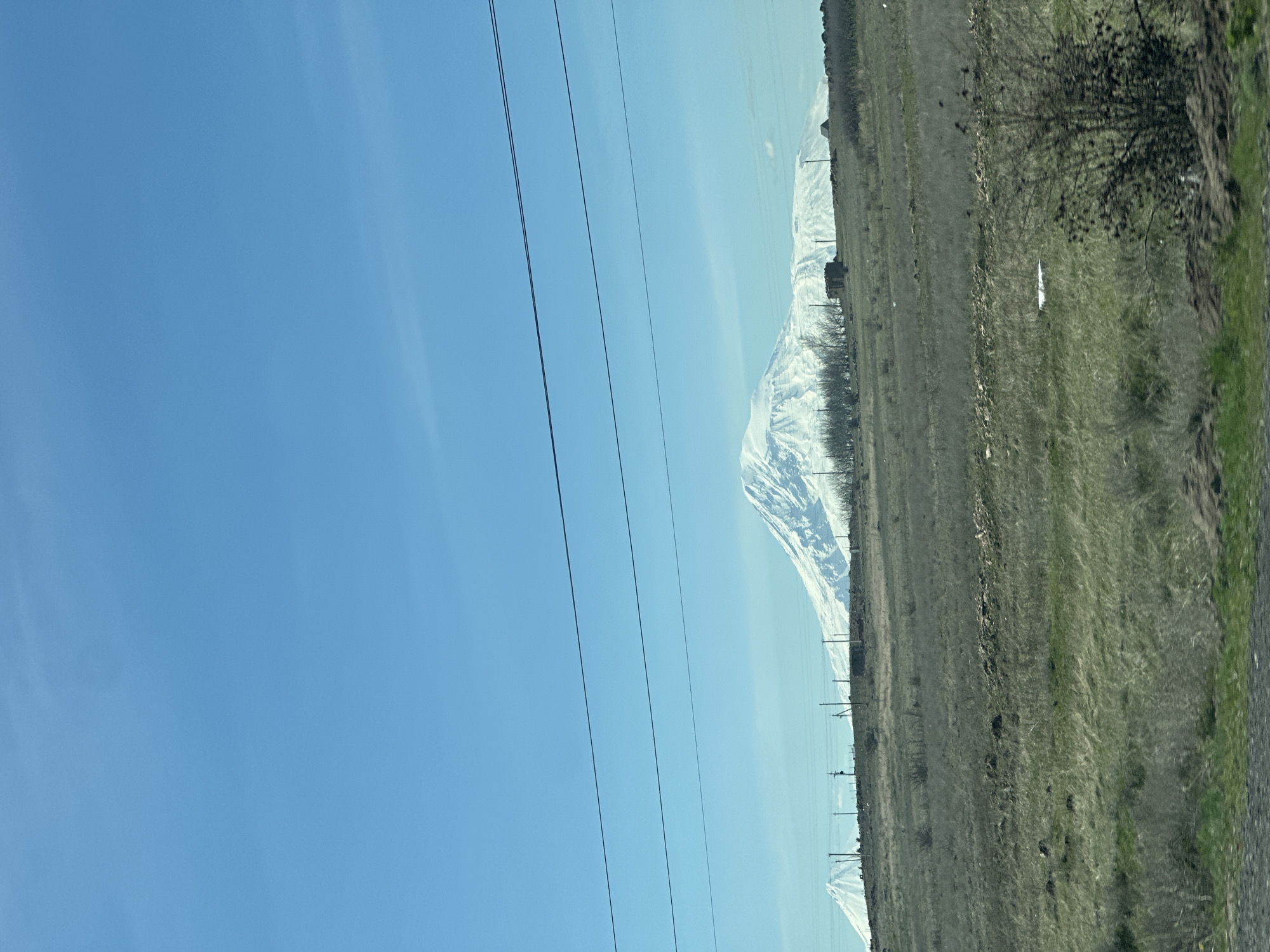 The image depicts a rural landscape under a clear, light blue sky. Electrical power lines stretch across the left side of the frame. To the right, a field with a mixture of green grass and brown vegetation is visible. Patches of snow are scattered in the field. In the distance, a snow-capped mountain range appears, partially obscured by a light haze. The image is taken from an oblique angle, suggesting it was captured from a moving vehicle.