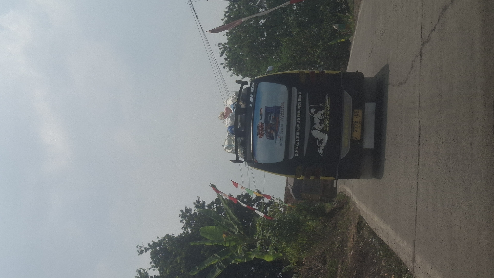 The image shows the rear of a black truck driving on a paved road in Sirnagalih, Indonesia. The truck has several items strapped to its roof, including bags and what appear to be rolls of fabric. A sticker on the back of the truck displays a graphic of a woman and text in Indonesian. The license plate is partially visible, reading "F 7776 WD". The road is flanked by trees and vegetation, with some Indonesian flags visible on poles to the left of the truck. The sky is overcast. The truck is positioned centrally in the frame, moving away from the viewer.