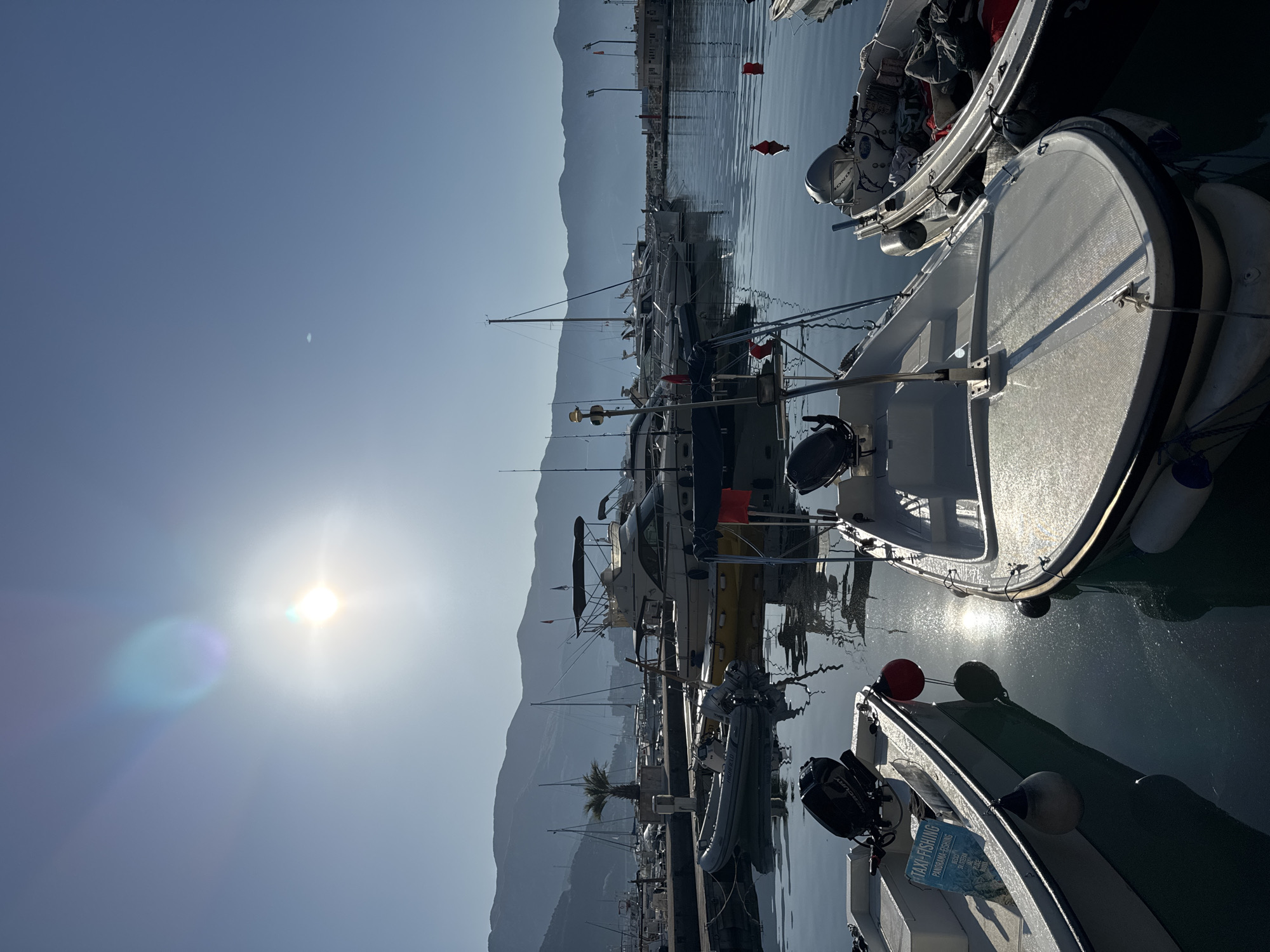 This image depicts a sunny day at a harbor in Budva, Montenegro. The sun shines brightly in the sky, casting a glare. Mountains are visible in the background. The harbor is full of boats, including dinghies. The reflections of the sun and the boats can be seen on the water.
