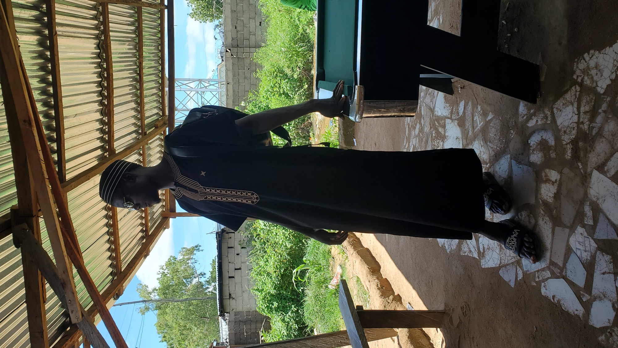 A full-body, slightly low-angle shot shows a Black man standing indoors, partially covered by a corrugated metal roof supported by wooden beams. He is wearing sunglasses, a black robe with gold trim around the neckline, and black Balenciaga slides. The man is facing right, with his right hand resting on the edge of a green-felt covered pool table. His left arm is bent, and a black backpack is visible on his back.
The floor is a mosaic of broken white and gray tiles with some dirt and debris. To the left of the man, a wooden bench with rough-hewn planks is visible. Behind the bench, there's a patch of dry, sandy ground. In the background, a brick wall topped with barbed wire separates the scene from lush green vegetation and trees under a bright blue sky with some clouds. In the distance, a metal tower-like structure can be seen. The overall impression is an outdoor or semi-outdoor recreational area, possibly a bar or community center, located in Jalingo, Nigeria.