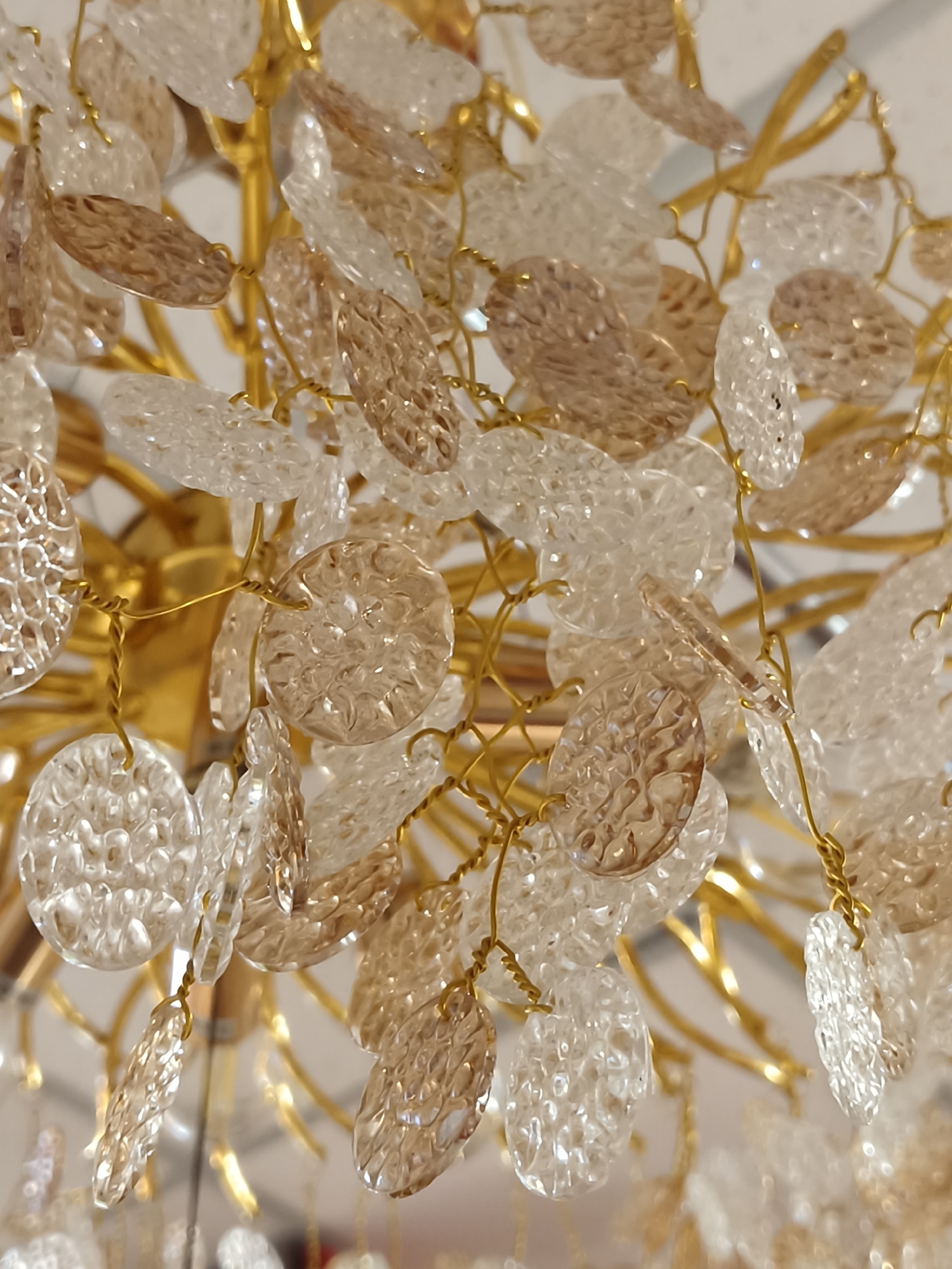 This close-up image shows a detail of a decorative chandelier, likely located indoors in Kryvyi Rih, Ukraine. The primary subjects are numerous small, textured, translucent pieces resembling petals or discs, which are suspended from a golden metallic structure. These decorative elements are presented in two main tones: clear and a warm, light amber or brown. Each piece features a textured surface with small, irregular bumps or facets that catch and reflect light, creating a sparkling effect.
They are individually attached by fine, twisted gold-colored wire to a more substantial golden metallic framework, which appears to consist of branches or arms that form the main body of the chandelier. The metallic parts are shiny and reflective. The background is softly blurred, emphasizing the intricate details of the foreground elements. No activity or event is taking place, as the image focuses on the static decorative object. There is no visible text, and the indoor lighting does not suggest a specific time of day or weather condition.