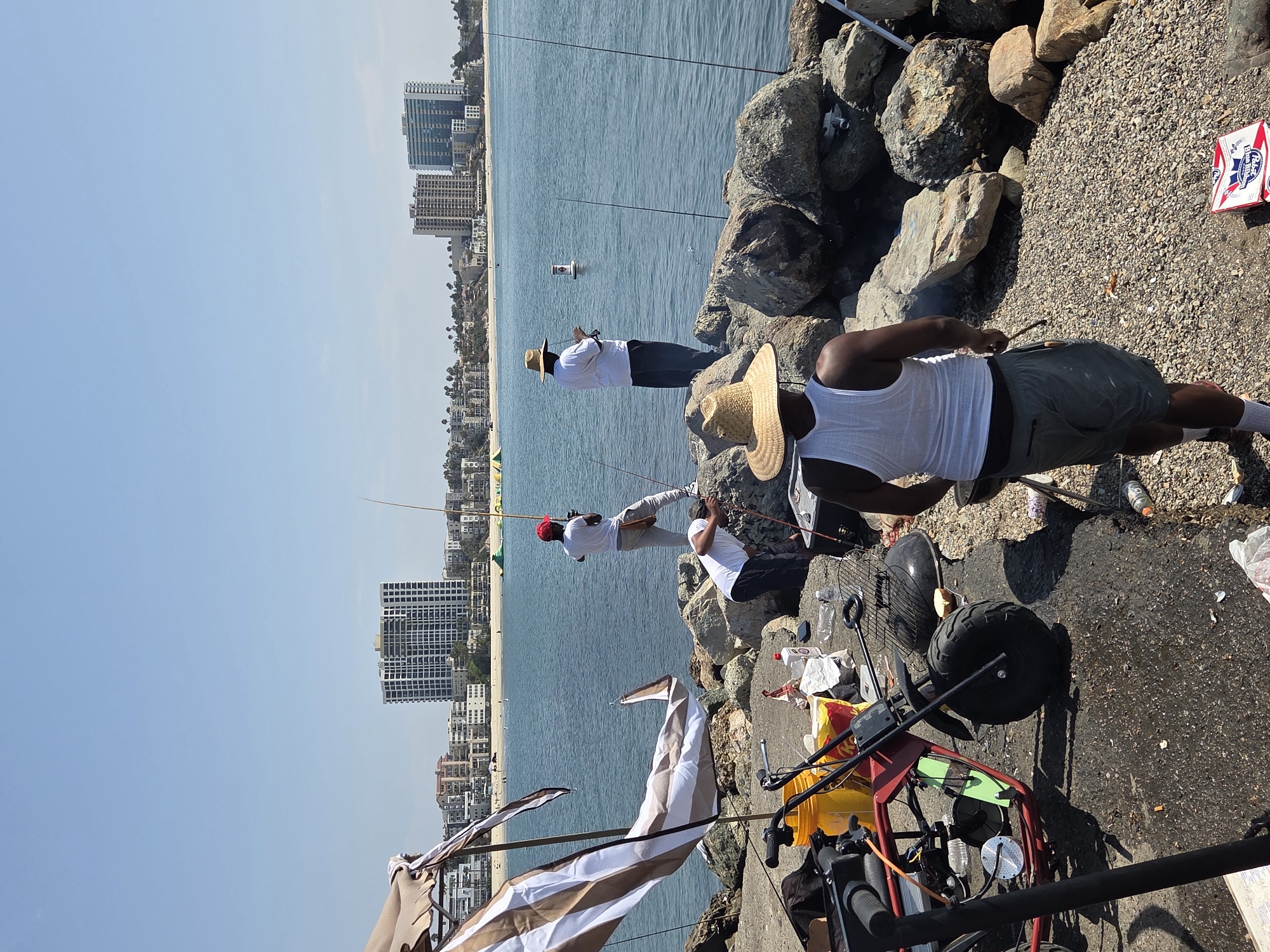 The image depicts a sunny day at the Long Beach harbor in California, with several people fishing from a rocky breakwater. In the background, high-rise buildings and a beach are visible, suggesting the urban environment of the city. The foreground shows a collection of fishing gear, a small red ATV-style vehicle, and a barbecue grill, indicating a recreational gathering. One individual, wearing a white tank top and a straw hat, is facing away from the camera and appears to be attending to the grill. Another person, also in a white shirt and straw hat, is fishing further out on the rocks. Two other individuals are fishing closer to the shore, one wearing a red cap and the other tending to fishing equipment. The water is calm and blue, reflecting the clear sky. The scene captures a leisurely activity of fishing and grilling by the waterfront in Long Beach.