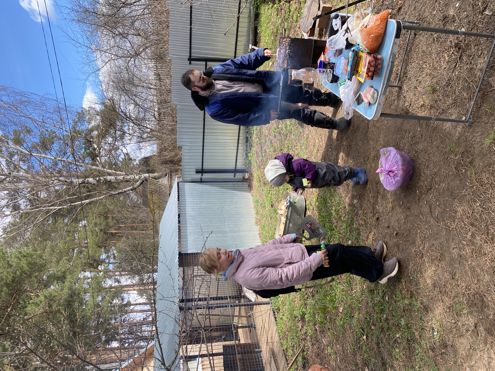 The image depicts an outdoor gathering near what appears to be a private residence. The location is near Chaikovsky, Russia, suggested by the architecture and landscape. Three individuals are present. A man in a blue jacket stands over a grill, tending to food, with smoke rising from the grill. A child in a purple jacket plays on the ground near the grill. A woman in a pink jacket sits nearby. A table is set up near the grill, covered with food and drinks. The area around them is dirt with some grass and scattered leaves. A metal fence encloses the property. Trees surround the area.