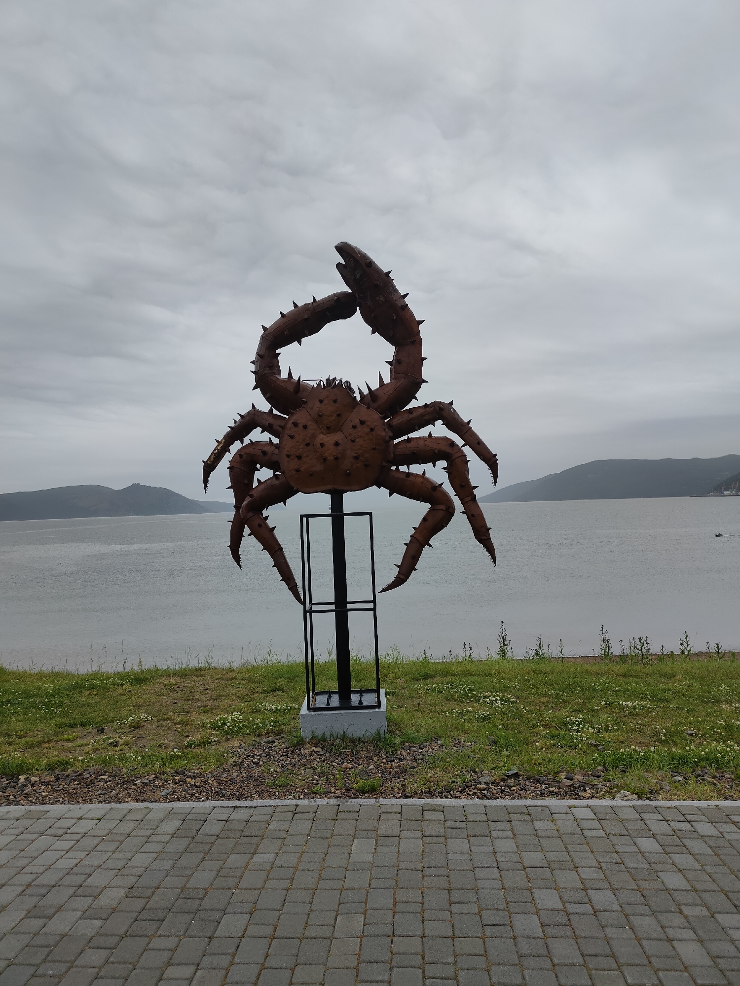An outdoor, eye-level, medium shot captures a large, rustic, brown metal sculpture of a crab situated in the city of Magadan, Russia, with the Sea of Okhotsk and distant hills forming the backdrop under a cloudy sky. The crab sculpture is centrally positioned, facing forward, and mounted on a dark metal post secured by a concrete base. Its legs are spread outwards, and its claws are raised. The sculpture is covered in sharp spikes and detailed with a textured surface. In the background, the calm sea stretches towards a hazy, mountainous coastline. A narrow strip of grass, dotted with small white flowers and pebbles, separates the paved walkway in the foreground from the sea. The walkway is paved with gray rectangular interlocking bricks, and a small boat is visible on the water in the distance.