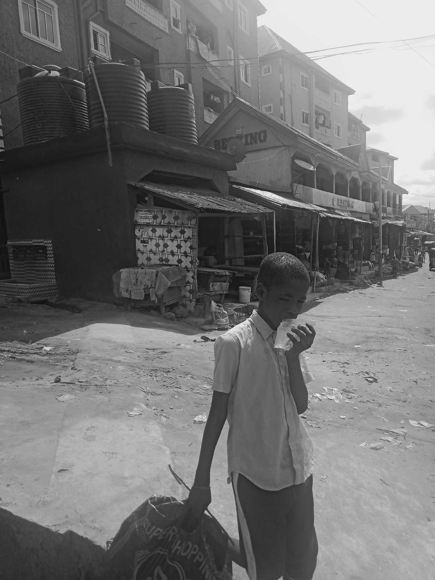 A black and white photograph captures a young person walking in a street in Onitsha, Nigeria. The person is seen holding a bag with the word "Super Shopping" printed on it. The person appears to be eating from a wrapper in their hand. The street is lined with buildings. There is litter on the ground. Water tanks are visible on top of a building to the left. In the background are shops and people. The lighting suggests it is daytime.