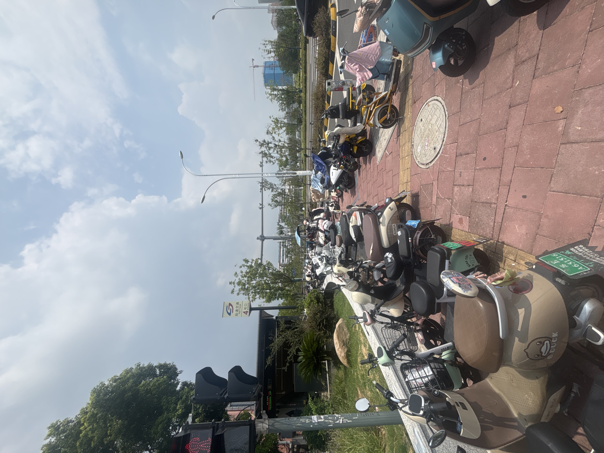 The image captures a wide shot of a street scene in Suzhou Industrial Park, China, on a bright, cloudy day. In the foreground, a row of parked electric scooters and motorbikes occupies a red brick sidewalk. The vehicles vary in color and design, with some covered by fabric. One scooter in the lower left corner features a "B. Duck" decal and a license plate that reads "A651516." The sidewalk is bordered by a low stone wall and a strip of green grass.
To the left, a traffic light with a red pedestrian signal is visible, along with a sign for "轨道交通" (Rail Transit) and "4号口" (Exit 4). Behind the scooters, a street with multiple lanes of traffic stretches into the background, with a black car passing on the right. Streetlights with modern, curved designs line the street. In the distance, trees and buildings are scattered across the landscape, with a construction crane and a partially completed blue building visible in the far background, indicating ongoing development. The sky is a mix of blue and white clouds. The scene is typical of an urban environment in China, with abundant electric two-wheelers for transportation and infrastructure development.
