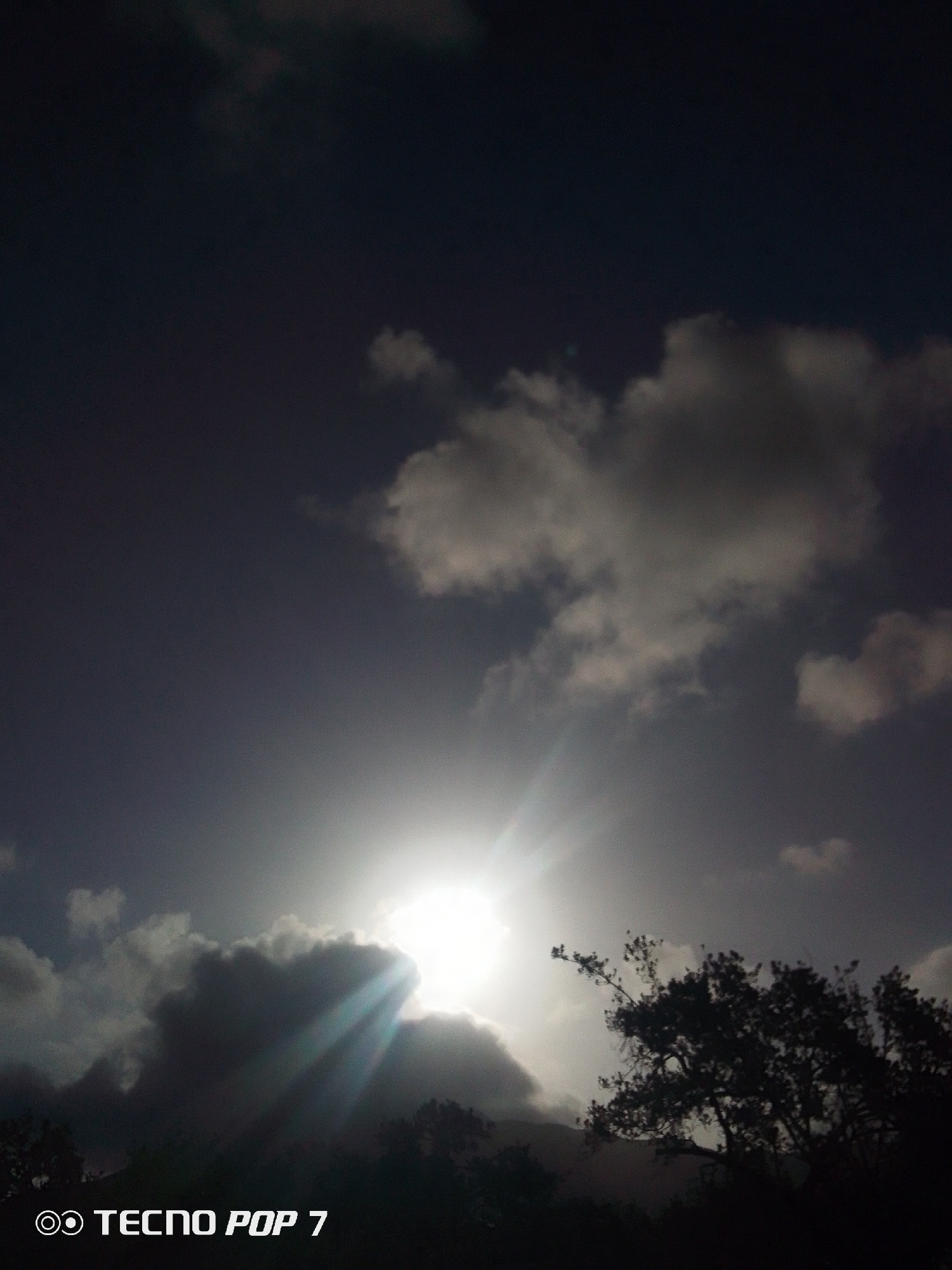 The image, taken in Pedro González, Venezuela, captures the sun obscured by clouds in the sky. The sun's rays peek through the clouds, creating beams of light. The sky is dark. Silhouetted trees and vegetation are visible in the foreground. The bottom of the image contains the text "TECNO POP 7."