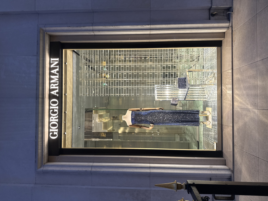 The image depicts the illuminated display window of a Giorgio Armani boutique, specifically located at 2, Avenue Montaigne in Paris, France. The scene is observed at dusk or night, indicated by the low ambient light contrasting with the brightly lit interior.
Within the display window, a single headless, armless mannequin stands centrally on a clear, elevated platform. The mannequin has wooden arms and is attired in a strapless, floor-length gown of dark blue fabric, heavily embellished with reflective sequins. Below the gown, the mannequin wears metallic silver, pointed-toe high heels.
To the right of the mannequin, a clear, geometric, multi-faceted structure, resembling a chair, serves as a display stand. Upon this structure, two dark-colored accessories are placed: a rectangular clutch with a metallic clasp to the left, and a matching, shimmering box-shaped clutch or small bag to the right. The background of the display features reflective surfaces and vertical, grid-like partitions, creating depth and reflecting various internal elements, including hints of gold-toned objects on shelves in the upper left.
The store's exterior facade is constructed of light-colored, possibly stone, material with prominent architectural molding surrounding the large display window. The "GIORGIO ARMANI" logo, illuminated in white, is prominently displayed above the window. The sidewalk in the foreground is paved with light-colored square tiles, reflecting the illumination from the display. A partial view of a black metal railing with a gold-colored, pointed finial is visible on the left side of the frame. No individuals are present outside the display window.