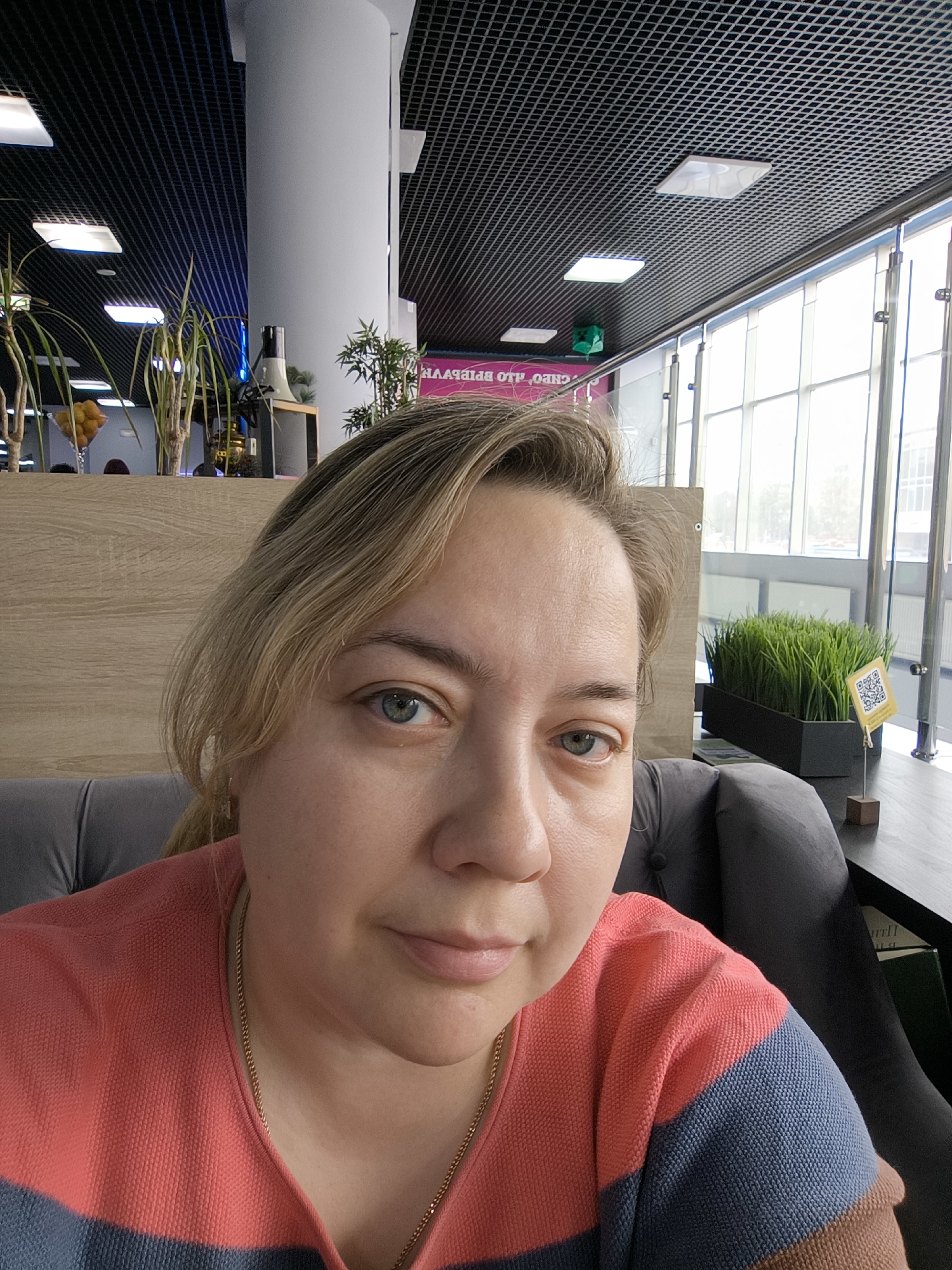 A close-up indoor shot features a woman with light blonde hair and blue eyes, wearing a pink and blue striped sweater and a gold necklace. She is seated in a grey upholstered chair.
Behind her is a modern interior space, likely a cafe or restaurant, with a dark, grid-patterned ceiling and bright, square-shaped light fixtures. There is a white cylindrical pillar, and a large window with vertical bars in the background. In front of the window, there are plants and a table with a small sign displaying a QR code. A wooden panel is behind the woman. There are other plants in the background, along with what appears to be a coffee maker and various other objects on a shelf. A sign in Cyrillic script is also visible in the distance. The location is in the city of Unknown, Unknown.
