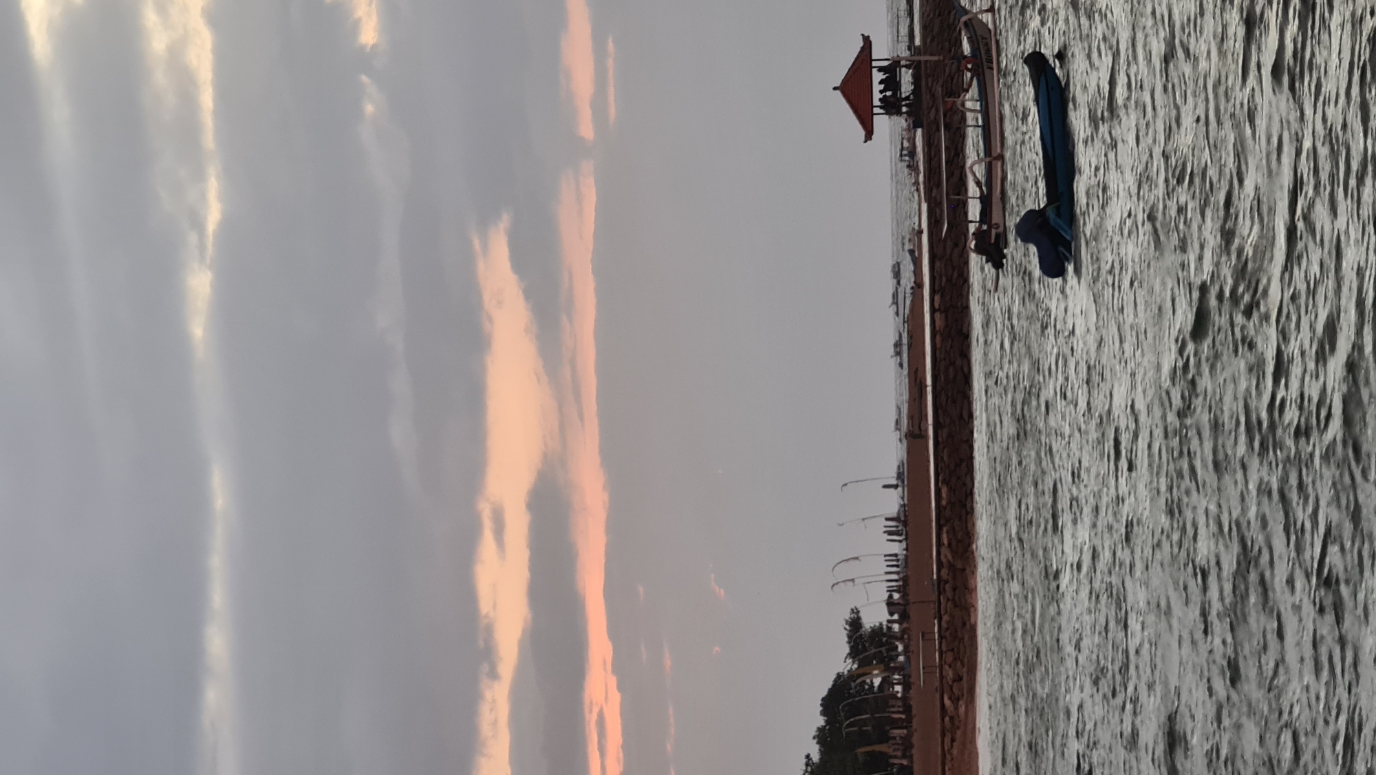 This media file depicts a waterfront scene in Denpasar, Indonesia, during what appears to be sunset. The sky is filled with dramatic clouds, painted with hues of gray, white, and soft pink and orange, suggesting the golden hour. The water, a prominent element, is textured and choppy, reflecting the muted light.
On the right side of the frame, a walkway or pier stretches along the water's edge. Along this walkway, several objects and structures are visible. A small, red-roofed pavilion with people gathered underneath is a focal point. Further along, a blue kayak or surfboard is propped up, hinting at water activities. There are also lampposts and some greenery, including trees, lining the walkway, suggesting a public area. The overall impression is of a peaceful, end-of-day atmosphere. The composition is oriented vertically, with the sky occupying the left half and the water and structures on the right.