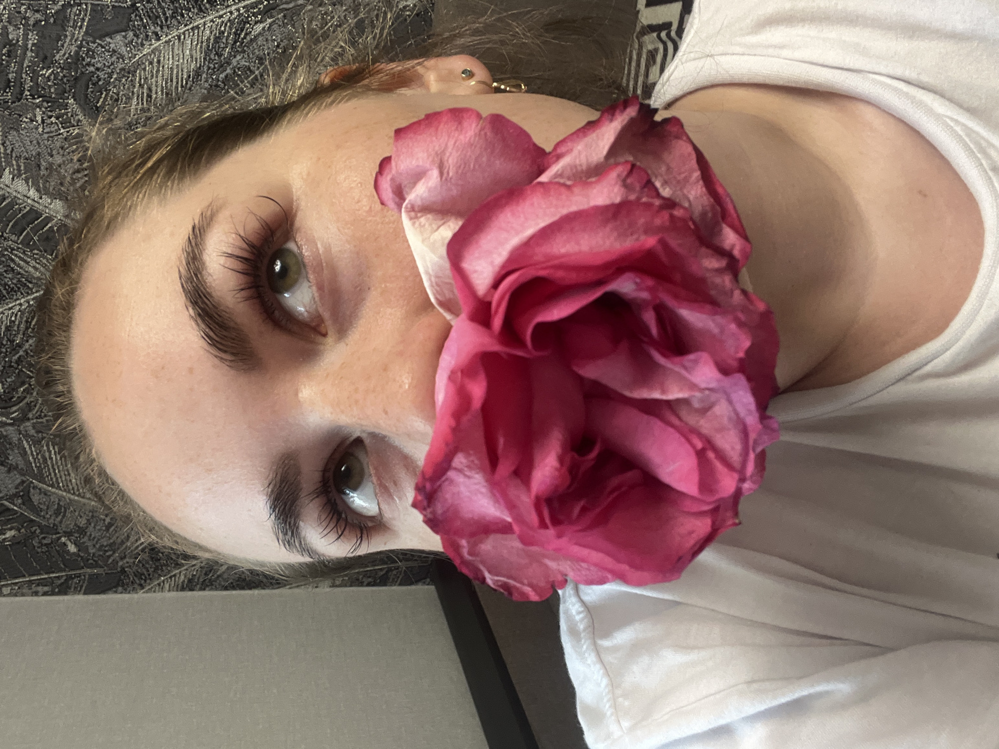 A woman's face is angled towards the camera, with a large pink rose obscuring part of her face. She has fair skin, light brown hair, and green eyes with dark, defined eyelashes. She wears a small gold earring. She is wearing a white shirt. The background is a dark gray textured surface. The location, Chaikovsky, Russia, is not evident in the image.