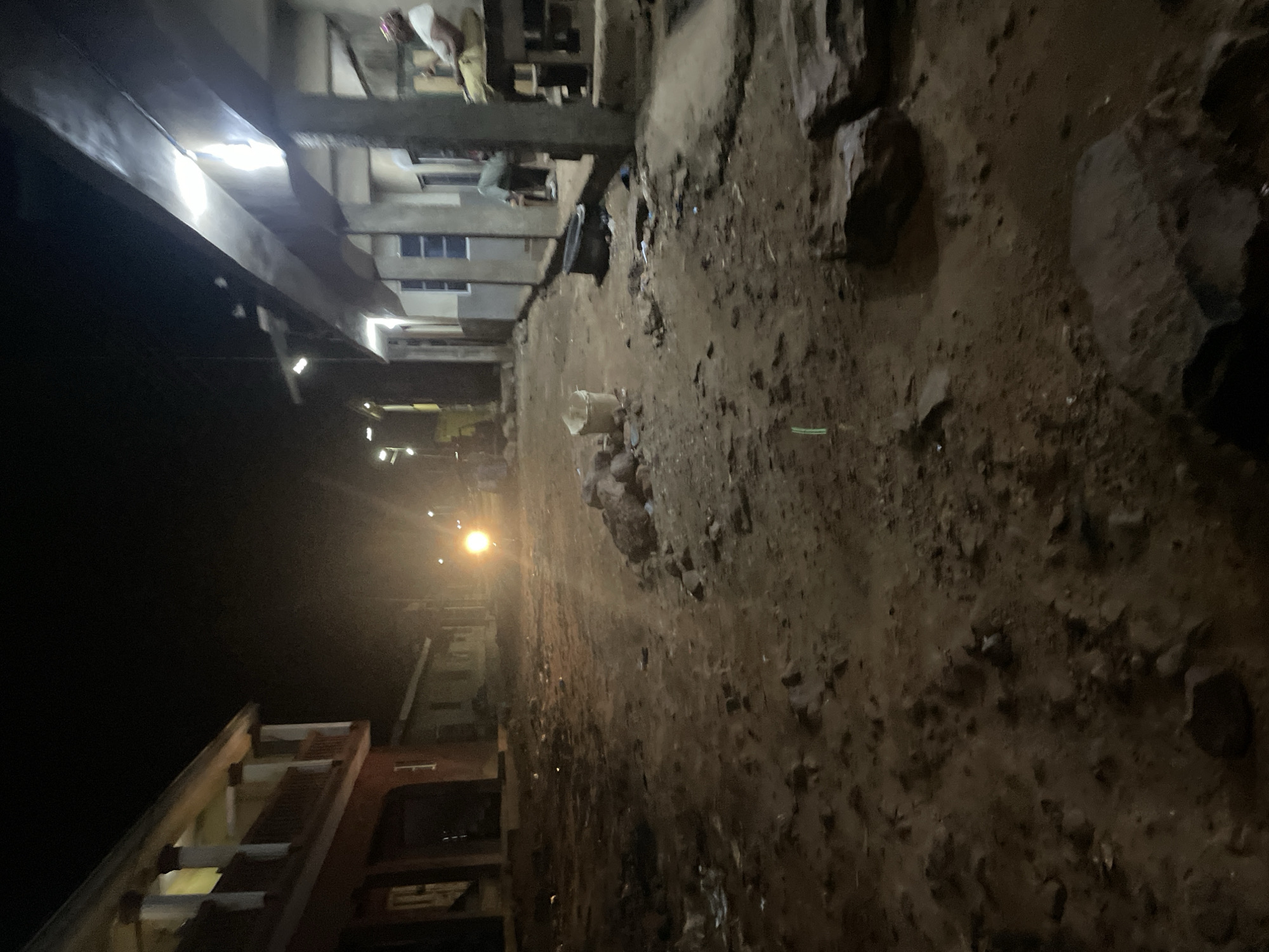 This image depicts a dimly lit street scene at night in Lokoja, Nigeria. The foreground is dominated by a dirt road littered with rocks and debris. On the left side of the street, a two-story building with a red facade and a balcony is visible. Further down the street, a vehicle's headlights create a bright glow. On the right side of the street, a series of connected buildings with covered walkways and windows are present. A person is seated on a table or platform under the covered walkway. The overall impression is of a sparsely populated, somewhat unkempt urban environment at night.