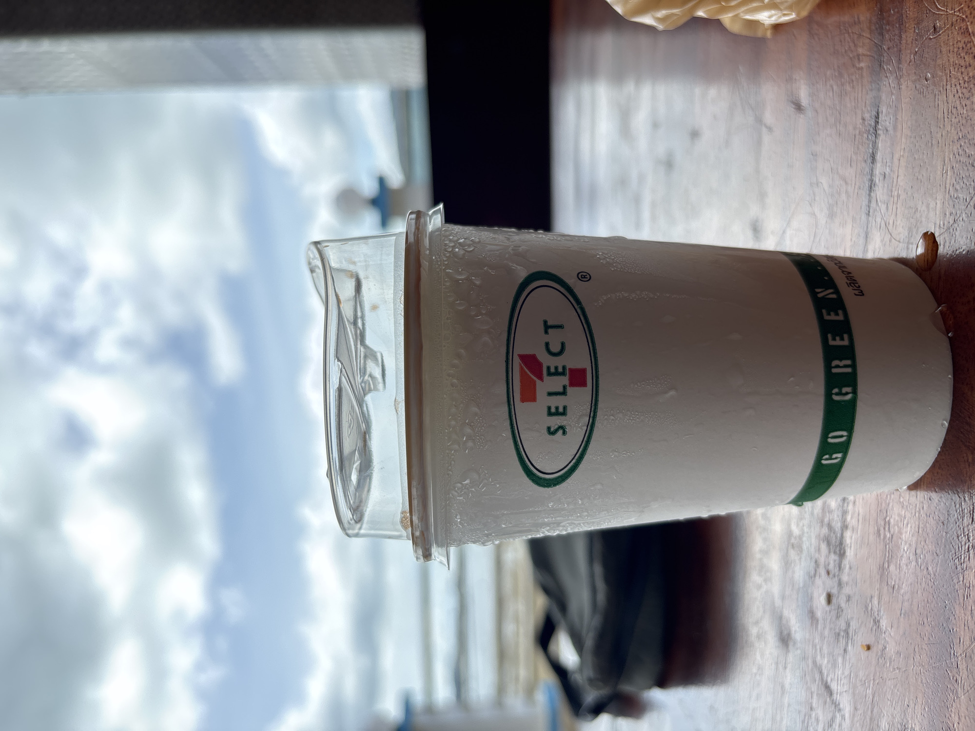 A condensation-covered, white paper cup with a clear plastic lid sits on a wooden table, its lid slightly ajar. The cup features a green and white oval logo with the number "7" and the word "SELECT" inside, and a green band near the bottom with the text "GO GREEN.". The background shows a blurry view of the sky with clouds, the ocean, and a dark object, possibly a bag, resting on the table. The scene is likely outdoors, given the natural light and background elements, possibly at a cafe or restaurant in Rawai, Thailand, overlooking the sea.