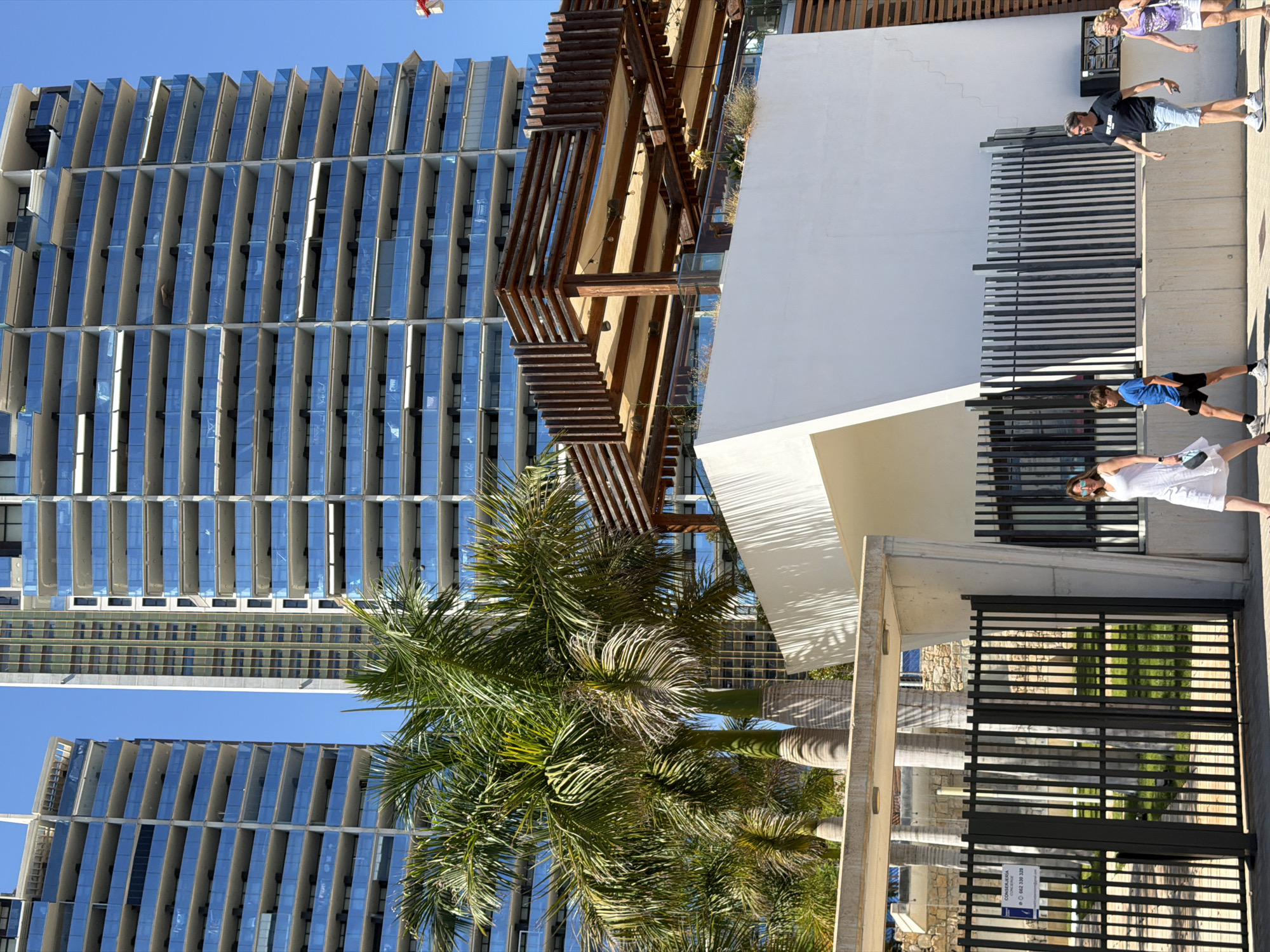The image depicts the front of a modern apartment complex in Benidorm, Spain, with palm trees in the foreground. Two tall, multi-story buildings with blue glass balconies dominate the background. In the middle ground, a white, angular structure with a wooden pergola roof is visible, along with some greenery. In the foreground, a black metal gate with a sign is present. Four people are walking on a paved area in front of the gate and buildings. From left to right, a woman in a white dress is walking, followed by a young boy in a blue shirt and black shorts, a man in a black t-shirt and blue shorts, and a woman in a purple and white top and a white skirt. The sky is clear and blue.