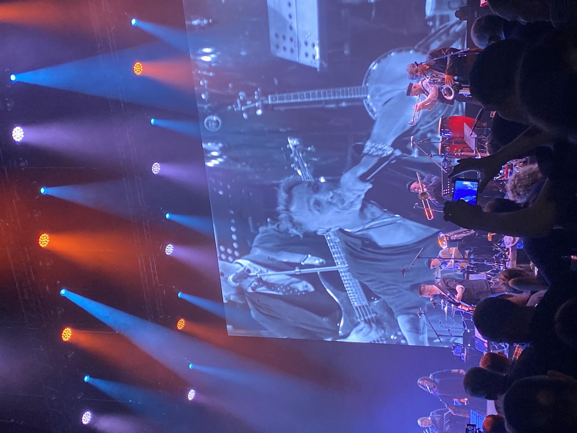 The image depicts a concert setting, possibly indoors, in the city of Yekaterinburg, Russia. A large screen displays a black and white close-up of a musician playing a bass guitar, surrounded by bandmates and musical equipment. The lower portion of the screen shows other band members, including a drummer. The foreground shows the heads and hands of audience members, with one person holding up a cell phone to record the event. Stage lighting consists of colorful beams, including blue, orange, and violet, illuminating the scene. The atmosphere appears energetic and crowded.