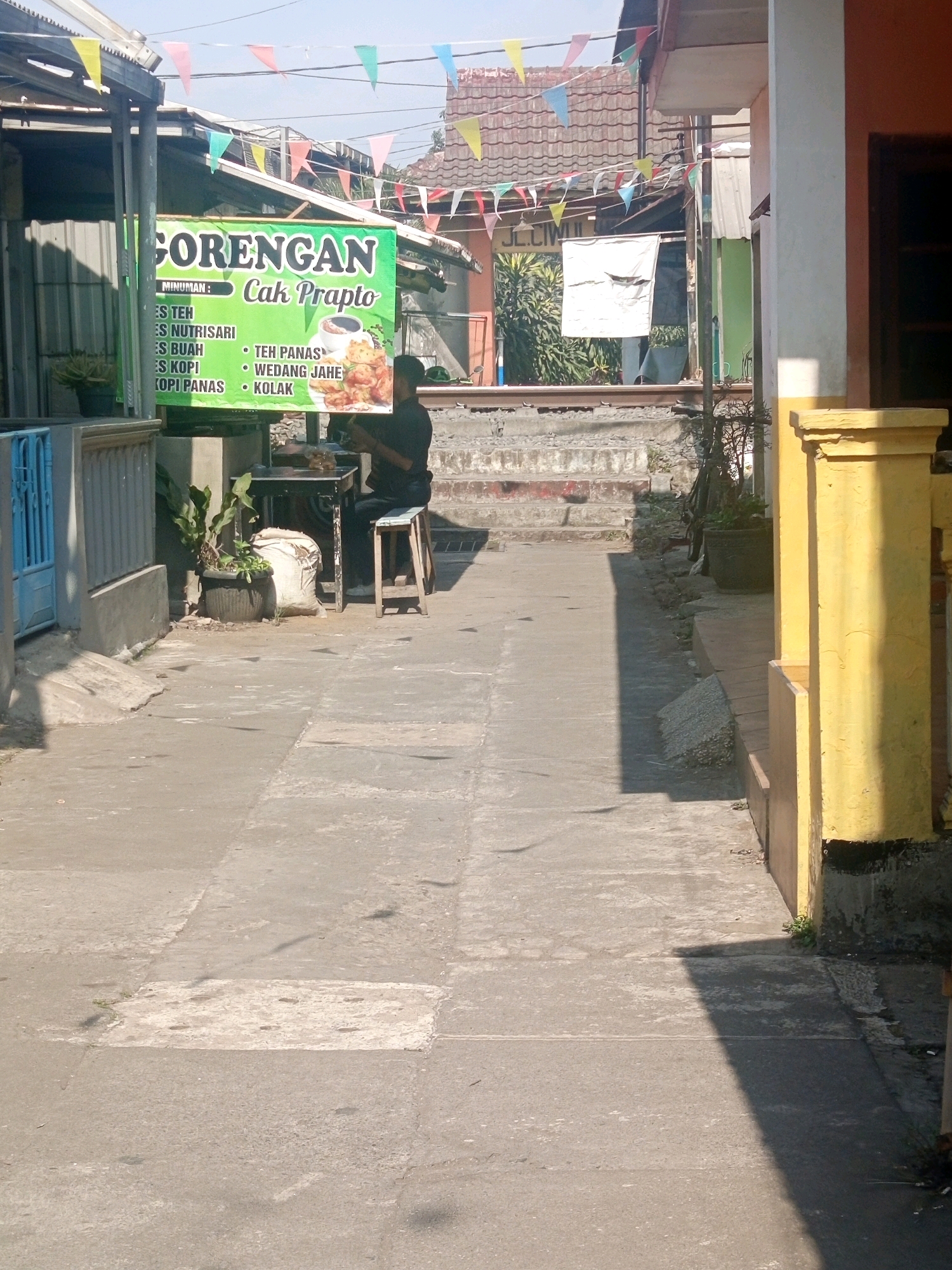 A narrow outdoor alleyway in Malang, Indonesia is depicted in the image. The alley is paved with concrete slabs of varying sizes and conditions. On the left, a blue metal gate and a grey concrete wall are visible. A green banner hangs from a structure, advertising "GORENGAN Cak Prapto" (Fried Snacks Cak Prapto) and listing various drinks and snacks such as "ES TEH" (iced tea), "ES NUTRISARI" (Nutrisari iced drink), "ES BUAH" (fruit ice), "ES KOPI" (iced coffee), "KOPI PANAS" (hot coffee), "WEDANG JAHE" (ginger drink), and "KOLAK" (a sweet dessert soup). Beneath the banner, a man in a dark shirt is seated on a wooden stool at a small black table, appearing to be serving customers or preparing food. A potted plant sits to his left, next to a large white sack. In the background, there are buildings with tiled roofs, some displaying festive triangular flags. The right side of the alley features a bright yellow pillar, part of a building structure. The scene is illuminated by daylight, casting shadows on the ground.