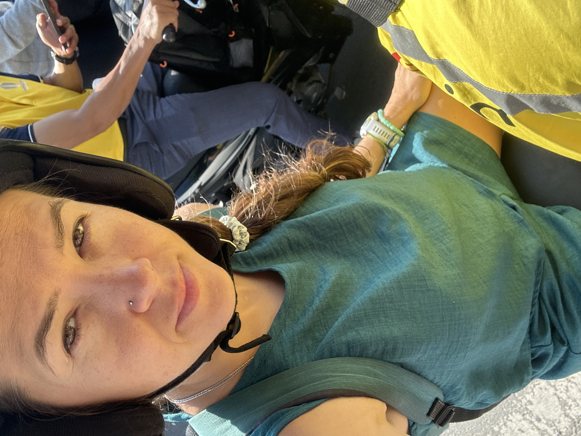 The image shows a woman wearing a black helmet, a teal sleeveless dress, and a silver necklace. She has a nose piercing and is wearing a white digital watch and green bracelets on her left wrist. She is seated on a motorcycle, with her right hand on the handlebar. Behind her, another person wearing a yellow shirt and blue pants is visible, holding a mobile phone. The background appears to be an outdoor urban environment, possibly in Nha Trang Ward, Vietnam, suggested by the presence of a motorcycle and attire commonly seen in Southeast Asia. The lighting indicates it is daytime.