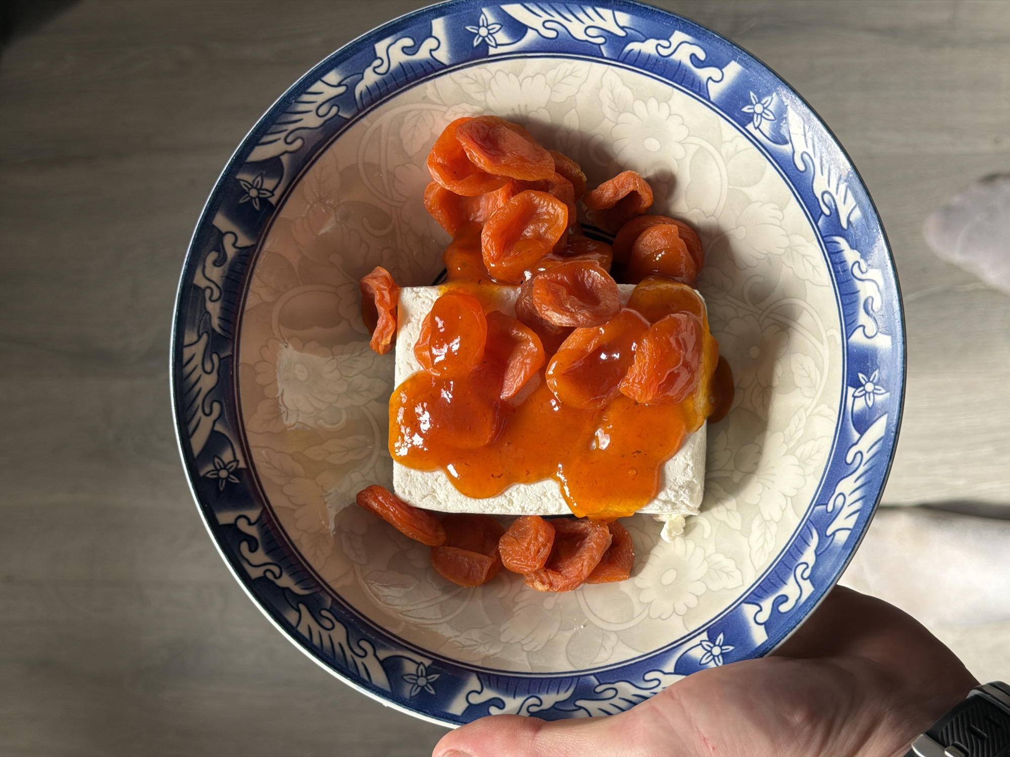 A high-angle close-up shot shows a bowl with a blue and white floral pattern filled with a white, square block (likely cheese or tofu) topped with dried apricots and some kind of orange sauce. The bowl is being held by a person. Part of their hand, wrist, and a watch are visible. The background is out of focus and appears to be a light-colored surface.
I have no information about the location.